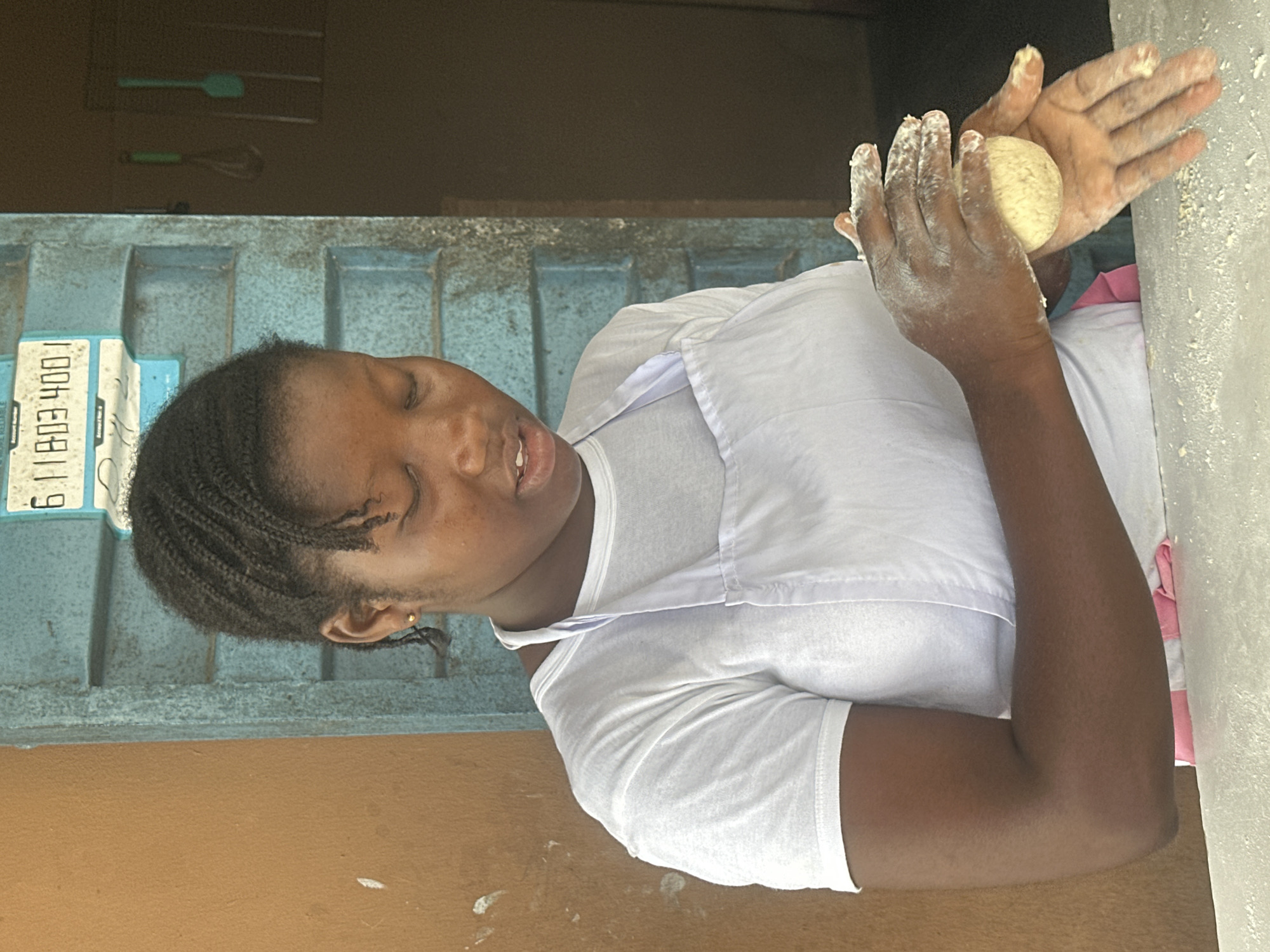A young woman with dark skin and braided hair is shown from the chest up, wearing a white t-shirt and a white apron. Her eyes are closed, and she appears to be concentrating on shaping a ball of dough with her flour-dusted hands. She is positioned in front of a weathered blue door with a sign displaying numbers in the upper right corner. To her left is a textured brown wall. In the background, on the door, are two kitchen utensils: a whisk and a spatula. The scene appears to be an outdoor or semi-outdoor food stall or kitchen, likely in the city of Badagry, Nigeria, given the context. Flour is visible on the counter in front of her and on her hands, indicating she is actively preparing food.