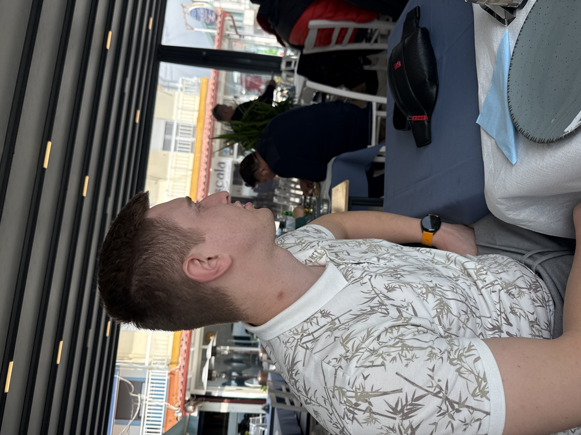 The image depicts a person with short brown hair and an orange wristwatch wearing a patterned white and tan shirt. They are seated at a table in what appears to be an outdoor dining area. Other people are seated at nearby tables in the background, with what seems to be a seaside view visible through the structures of the dining area. The outside scene includes buildings with colorful facades. This scene is located in Fanari, Greece.