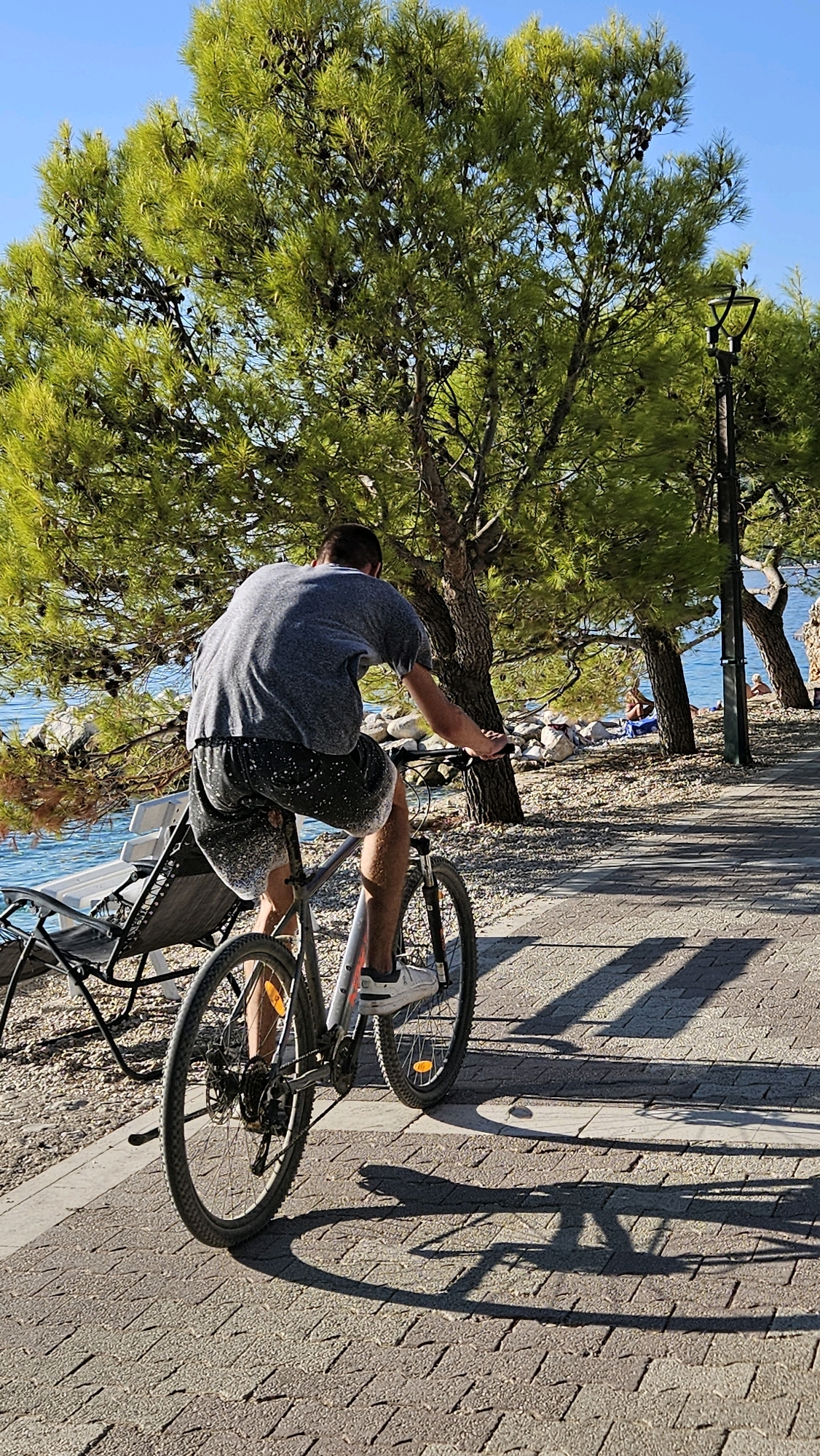 A person is riding a bicycle on a paved walkway next to a body of water. The person is wearing a grey t-shirt and patterned shorts. In the background, there are trees with green foliage, a beach with people, and a street lamp. The walkway is made of brick pavers, and shadows from the trees and the bicycle are cast across it. The water appears calm and blue, suggesting a sunny day. The scene is likely located in a coastal area, possibly Općina Tučepi, Croatia, given the presence of pine trees and a beach.
