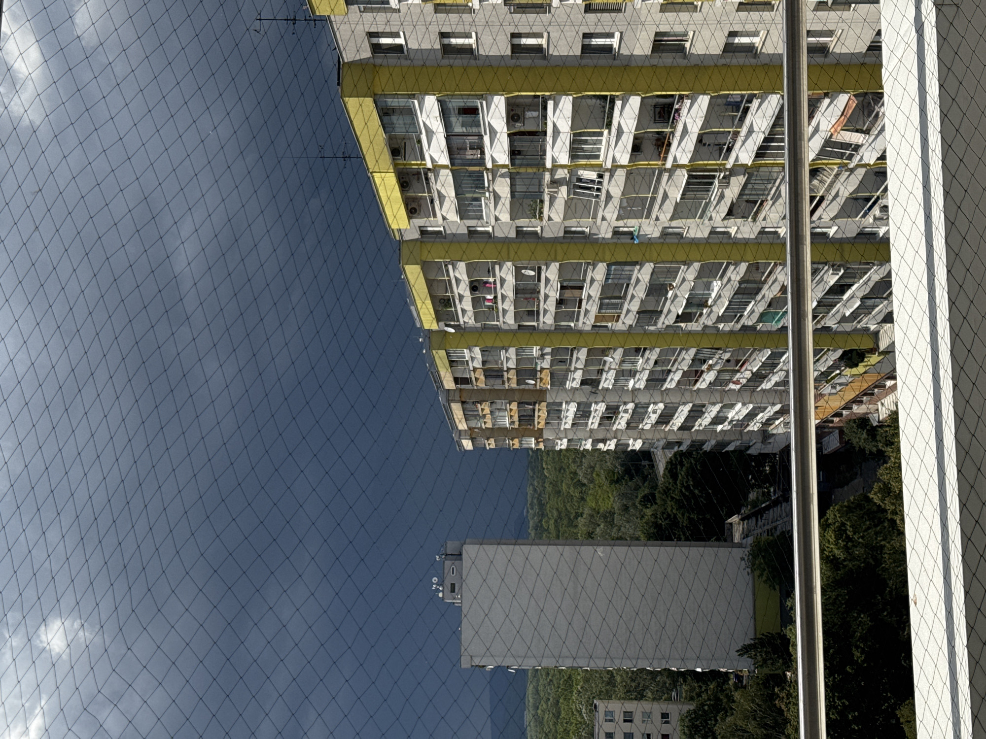 This is a vertical, eye-level shot looking out over a city skyline. The foreground shows a metal railing and a net. The mid-ground features two apartment buildings. The building on the right is tall and narrow with a grey facade and yellow accents along the balconies. The building on the left is wider with a light grey facade and a flat roof. Both buildings are densely populated with balconies and windows. Behind the buildings, a dense green forest stretches out towards the horizon. The sky above is a dark, stormy grey with hints of white clouds. The overall impression is of an urban environment in potentially inclement weather, with a view of natural surroundings. This is a view from a residential building in Bratislava, Slovakia.