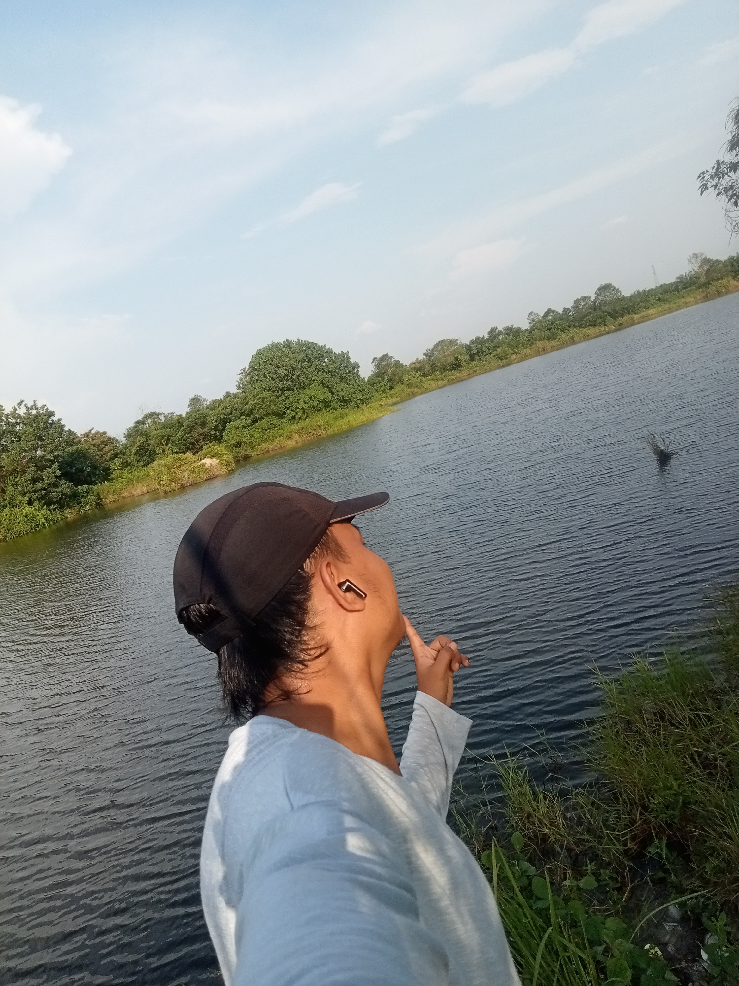 A person wearing a black baseball cap and a light grey long-sleeved shirt stands on the grassy bank of a large body of water, looking out across the water. The person has their right hand raised, with index finger pointing upwards. A black wireless earbud is visible in their right ear. The water is dark and rippled, with a line of trees and lush green vegetation on the opposite bank. The sky above is a pale blue with scattered white clouds. The scene appears to be set in Kualu, Indonesia.