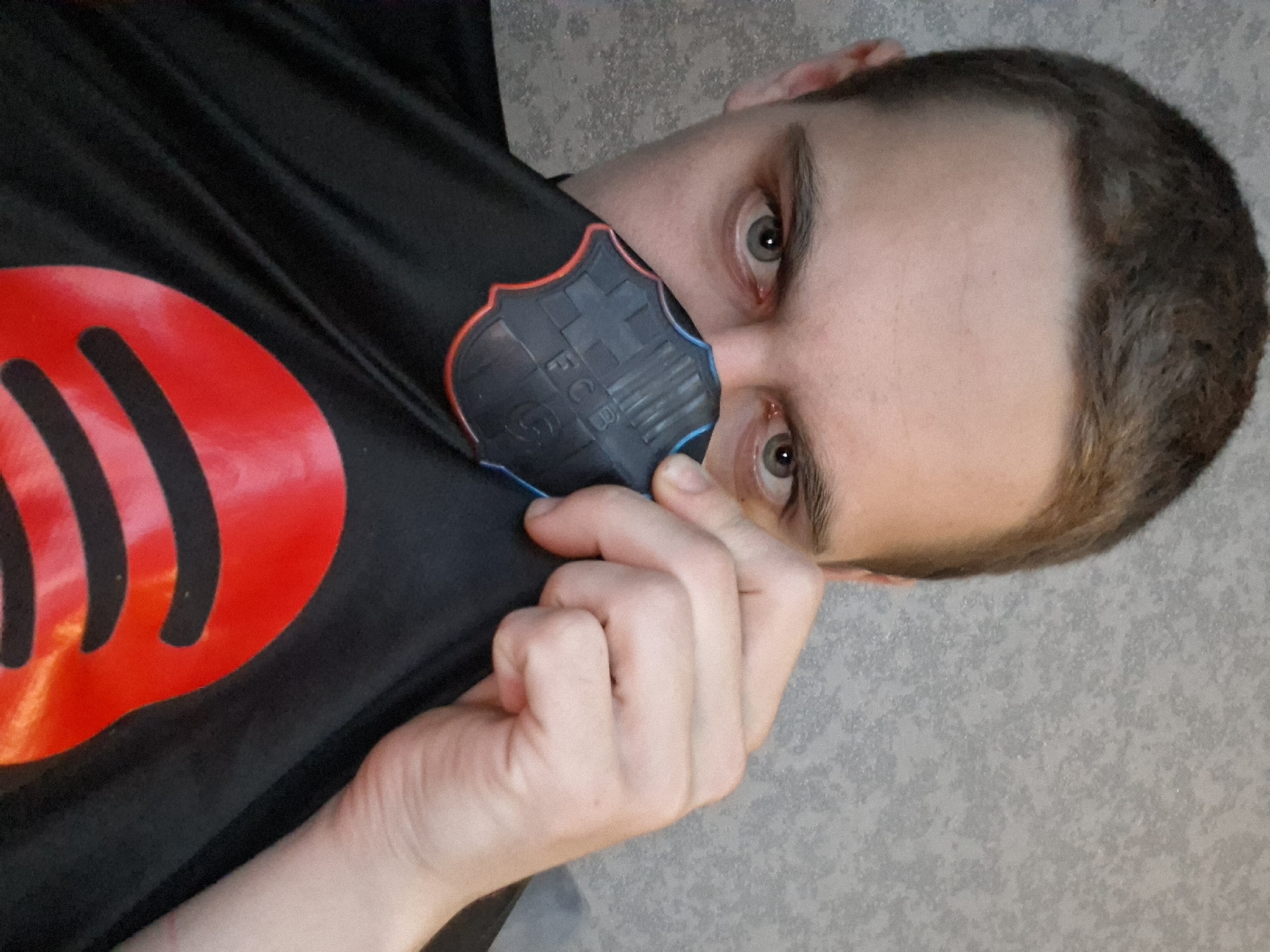 A man is holding a black garment with a red Spotify logo and a black FC Barcelona logo near his face. The background is a gray textured wall. The location is Unknown, Unknown.