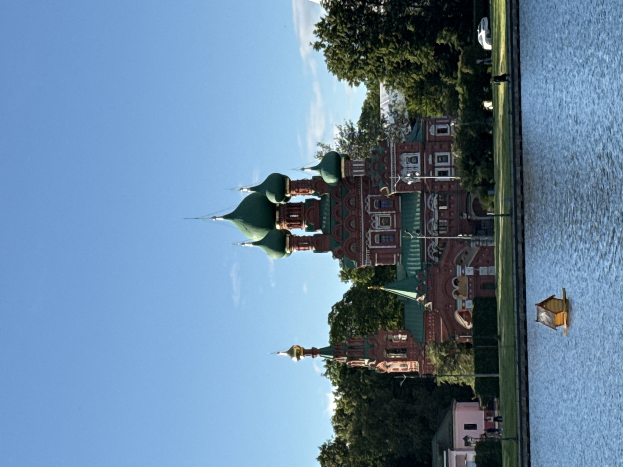 This media file captures a scene of the Sretensky Monastery in Moscow, Russia, on a clear, bright day. The image is oriented vertically, with the sky occupying the upper portion. The monastery itself is the dominant subject, with its distinctive red brick architecture and ornate green domes. Several onion-shaped domes with pointed spires are visible, glinting in the sunlight. The monastery is surrounded by lush green trees, suggesting a park-like setting.
To the right of the monastery is a large body of water, possibly a pond or river, its surface rippled. A small, yellow wooden structure, resembling a decorative birdhouse or a small boat, floats on the water. The time of day appears to be late afternoon, given the warm lighting and long shadows cast by the trees. The weather is fair, with a clear blue sky and a few wispy white clouds.
While the primary focus is the monastery and its surroundings, there are hints of human presence. In the distance, a white car is parked on a path adjacent to the trees, and a few figures can be seen walking or standing near the water's edge. The overall mood is serene and picturesque, highlighting the architectural beauty of the monastery against the backdrop of nature. No visible text is present in the image.