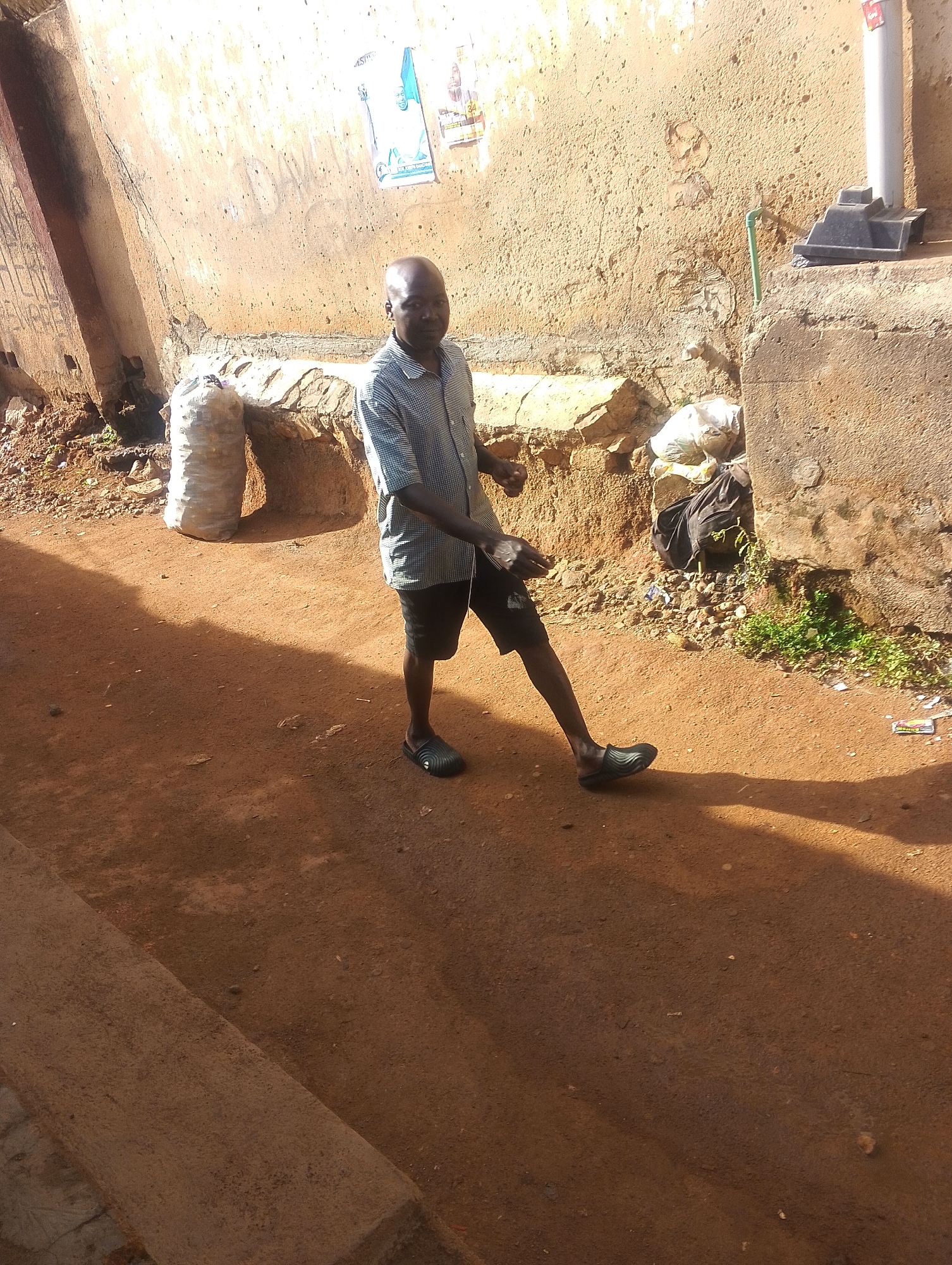 A man walks down a narrow, red dirt lane in Jos, Nigeria. He is bald and has dark skin, wearing a short-sleeved button-down shirt and dark shorts. He wears dark, rubber slip-on shoes. He faces the camera, and his left arm is out to the side, as if walking. A concrete wall is to his right, and on the left is a raised curb. Behind the man are two small openings in the wall. One opening contains a sack, and the other appears to have trash in it. A drainpipe is mounted on the wall. The background walls are cracked and worn. Posters are mounted on the wall near the drainpipe.