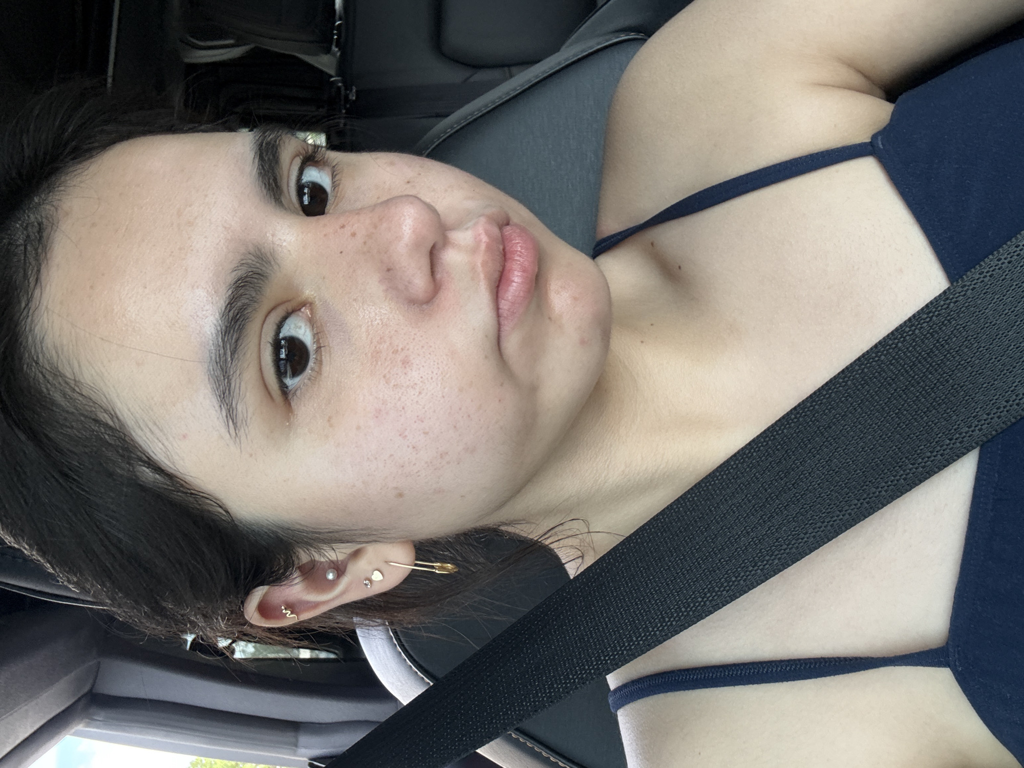 A young woman is in a vehicle, as indicated by the safety belt across her chest and the interior view of the vehicle. She is fair-skinned and has dark hair, with multiple piercings in her left ear. She is wearing a blue top with thin straps. The interior of the vehicle includes a dark grey seat and trim, suggesting it is a modern car. The woman appears to be tilting her head, and the camera angle is from above, possibly a selfie. It is not possible to determine the location of the vehicle as being in the city of Austin, United States of America, due to lack of environmental details.