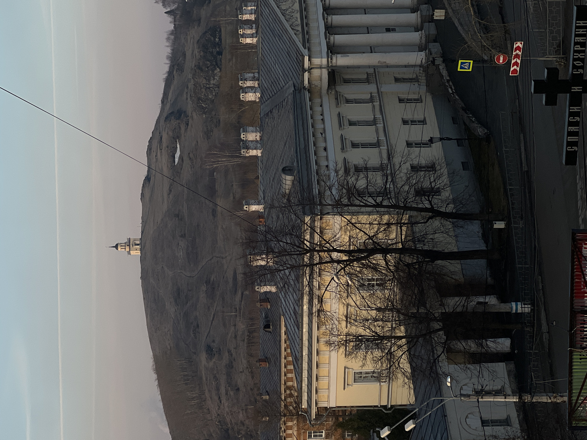 The image shows a view of Nizhny Tagil, Russia. In the foreground, there's a multi-story building with a light yellow facade and a grey roof. Bare tree branches obscure parts of the building. Street signs and a cross monument are visible on the right. In the background, a large, bare hill rises, with a small structure resembling a watchtower or church at its summit. The sky is pale, suggesting either dawn or dusk, and contrails from aircraft are visible. The hill is likely Fox Mountain, a well-known landmark in Nizhny Tagil.