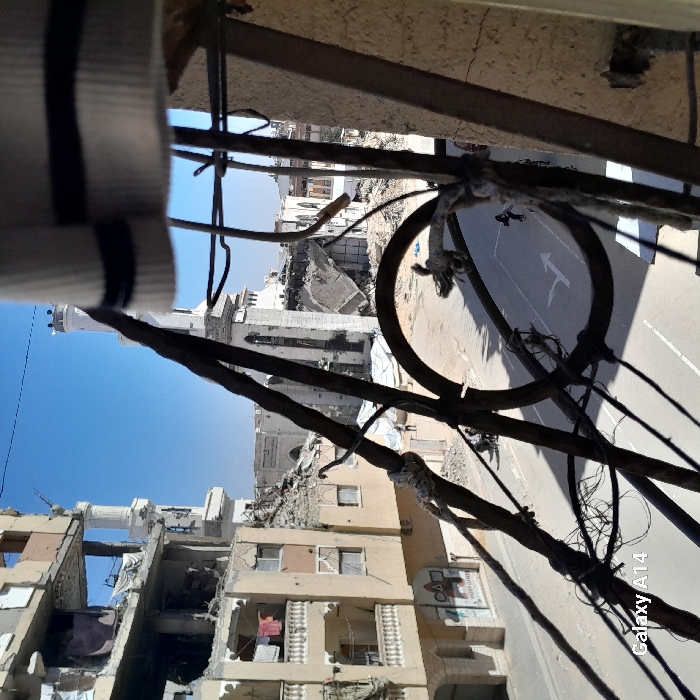 The image portrays an urban scene in Khan Yunis, Palestinian Territory, observed from an elevated perspective through a foreground partially obscured by dark, rusted metal bars, wires, and a white fabric item with black stripes at the top right.
On the left, a multi-story residential building, light-colored, displays extensive structural damage, particularly on its upper floors where walls are missing and rebar is exposed amidst rubble. Clothes, including red, pink, and white items, are visible hanging on a line from a lower-level balcony of this building. A sign with Arabic script and a red and white triangular logo is affixed to the building's street-level facade.
In the mid-ground and background, a large white mosque-like structure with at least two minarets is prominently featured, exhibiting significant damage, including sections of its roof and walls collapsed into surrounding debris piles. A large white tarp or tent is situated near the base of the mosque. The street below is paved, marked with a white arrow indicating a right turn. Two individuals are visible on the street: one near the center, appearing to walk or push an object, and another further to the right. The sky is clear and blue, indicating daytime conditions.