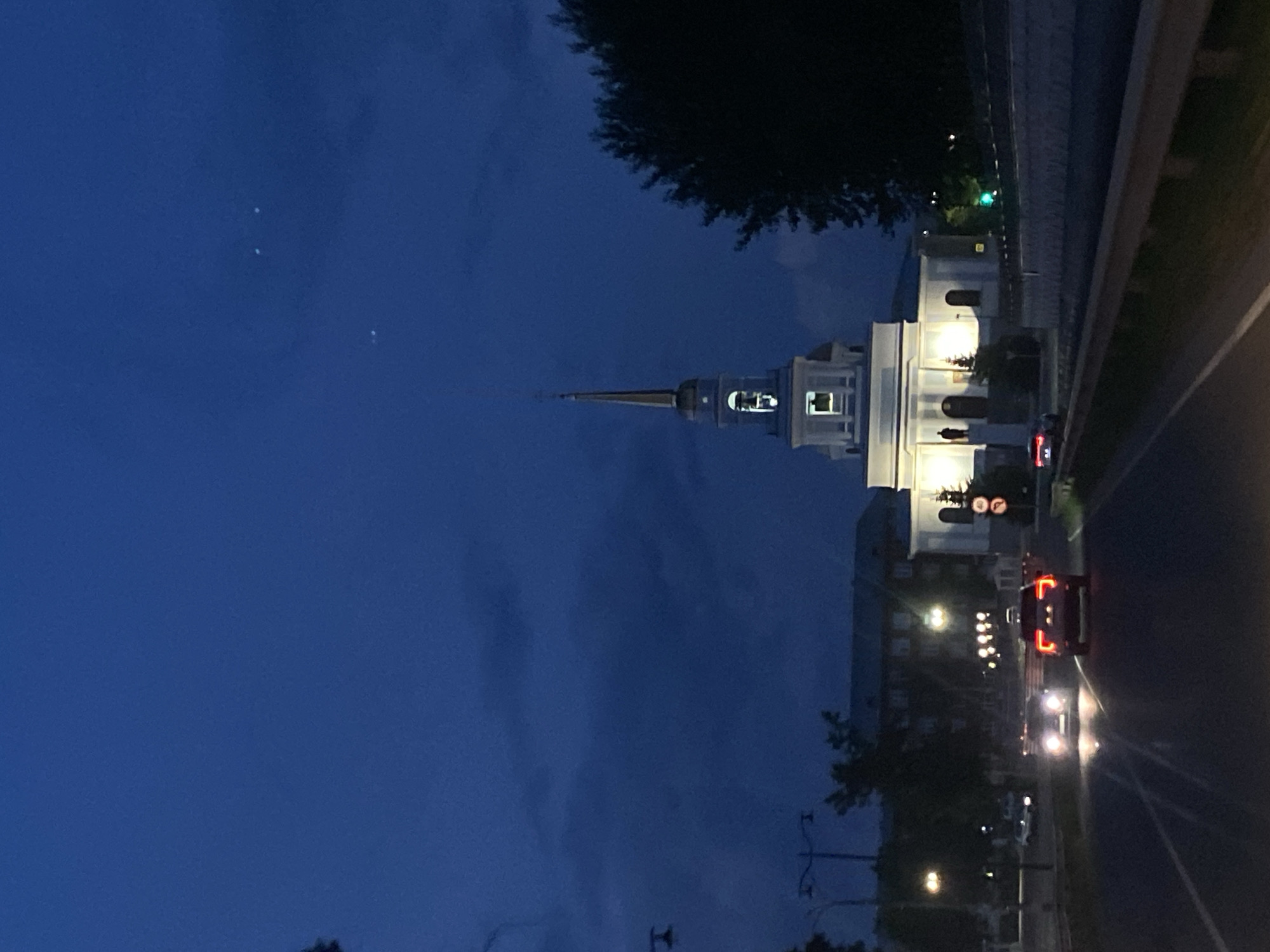 The image depicts a view of a church and surrounding cityscape in Votkinsk, Russia, during twilight or early night. The prominent structure is a white church with a tall, slender spire topped with a golden finial. The church has illuminated windows, and a bell is visible in an arched opening near the top of the main body of the building. In front of the church, a dark, silhouetted statue stands on a pedestal.
The scene includes a road in the foreground with several cars. Two cars are driving away from the viewer, their taillights illuminated. Another car is approaching the viewer, its headlights on. The road is bordered by a metal guardrail. In the background, there are buildings, streetlights casting pools of light, and trees. The sky is a deep blue, with some wispy clouds and a few faint celestial points visible. The overall atmosphere is quiet and serene, illuminated by artificial light against the darkening sky.