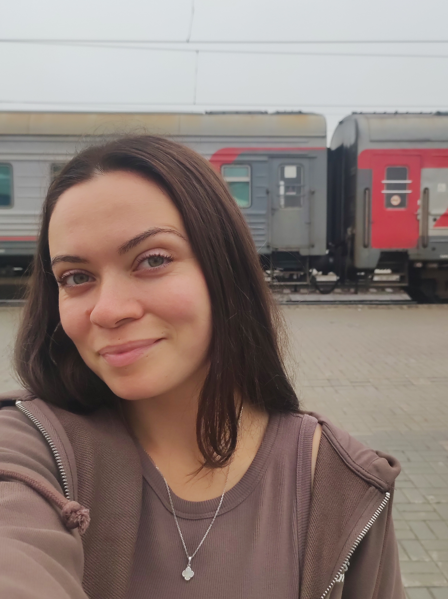 A young woman with long, dark hair and light green eyes is captured in a selfie at a train station in Kirov, Russia. She is smiling softly at the camera, her head tilted slightly. She is wearing a brown zip-up hoodie over a mauve ribbed tank top, adorned with a silver necklace featuring a clover-shaped pendant. Behind her, a train with gray and red carriages is visible, stopped at the platform. The platform is paved with light-colored tiles, and the sky above is overcast.