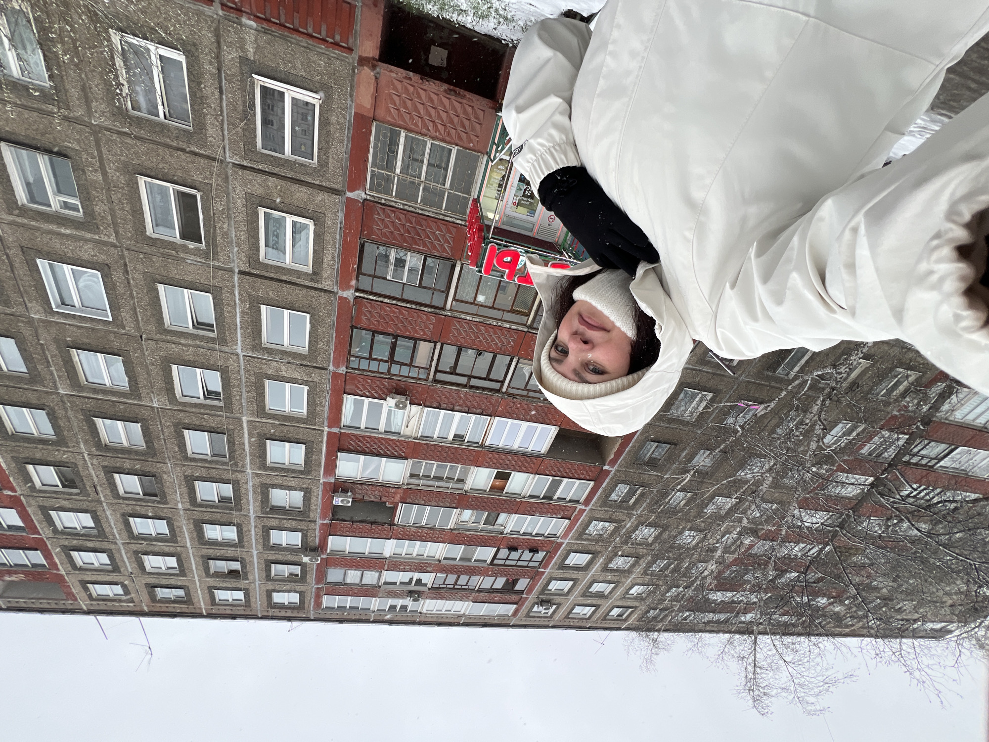 A young woman is taking a selfie outdoors in an urban setting during winter in Perm, Russia. She is positioned on the right side of the frame, wearing a warm white hooded winter jacket, a white knitted hat, and black gloves. Her face is visible, looking directly at the camera with light-colored eyes and a neutral expression.
The background features two large, multi-story apartment buildings. The building on the left has a grey, textured facade, likely made of concrete panels, with many square windows arranged in a grid. To its right and slightly further back, a second reddish-brown apartment building stands, characterized by a repeating patterned facade and numerous windows, some fitted with security bars and external air conditioning units.
The scene depicts a cold, overcast winter day with visible light snowfall. Bare tree branches, dusted with snow, are scattered throughout the midground, and the ground beneath the buildings appears snow-covered. The architecture of the buildings is typical of urban residential areas in Russia, suggesting a post-Soviet era construction.
A prominent red sign with white Cyrillic text is visible on the reddish-brown building. While partially obscured and possibly mirrored, the large letter "Я" (Ya) is discernible. Smaller text appears to include "24 ЧАСА" (24 HOURS) and the word "DRY," suggesting a commercial establishment such as a dry cleaner or laundry. The image conveys a sense of a brisk winter day in a city environment.