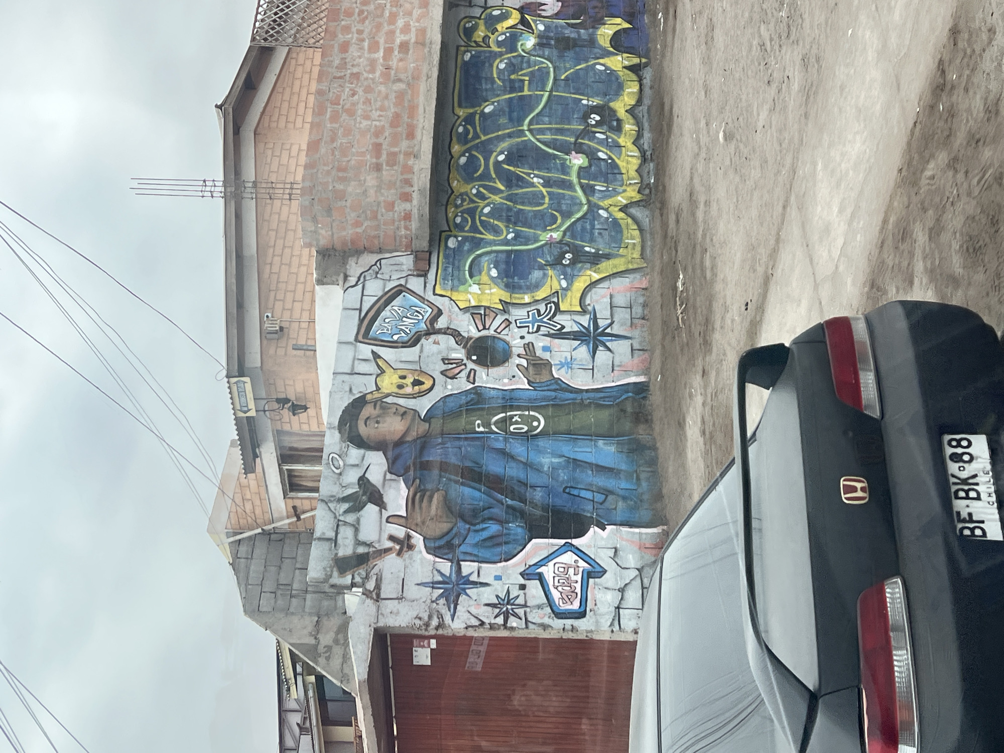 Arica, Chile, street scene shows a black Honda automobile, rear view, partially blocking a mural on a brick wall. The mural depicts a young man in a blue jacket, surrounded by cartoonish figures and graffiti-style lettering. The wall is adjacent to a building with both brick and concrete block construction. Overhead electrical wires are visible against a cloudy sky. The car's license plate reads "BF BK 88, CHILE".