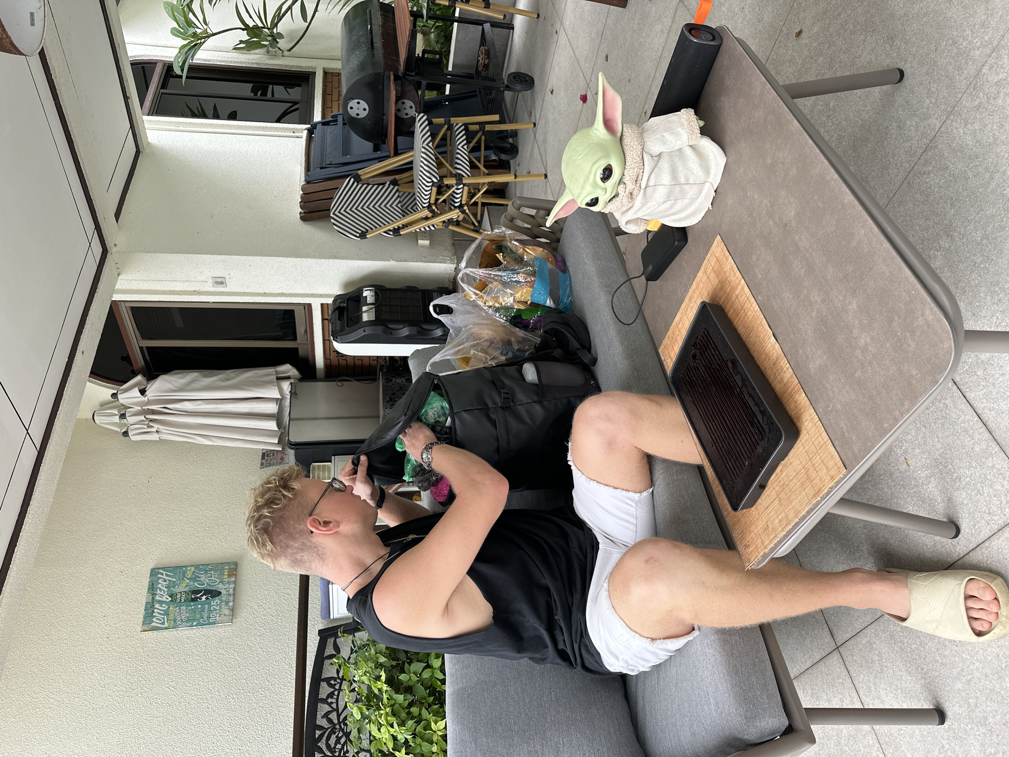 A blond man wearing a black tank top and white shorts is sitting on a gray outdoor sofa and rummaging through a black duffel bag. He has on glasses and several necklaces. To his right, on a gray table, sits a small Grogu plush toy, a black portable charger, and a black soundbar. A portable grill with a brown bamboo mat is in front of the Grogu toy on the table. Behind the man, there are several plastic bags filled with items. Further back, there are grills, chairs, and a white portable air conditioner unit. The scene appears to be an outdoor patio or porch area.