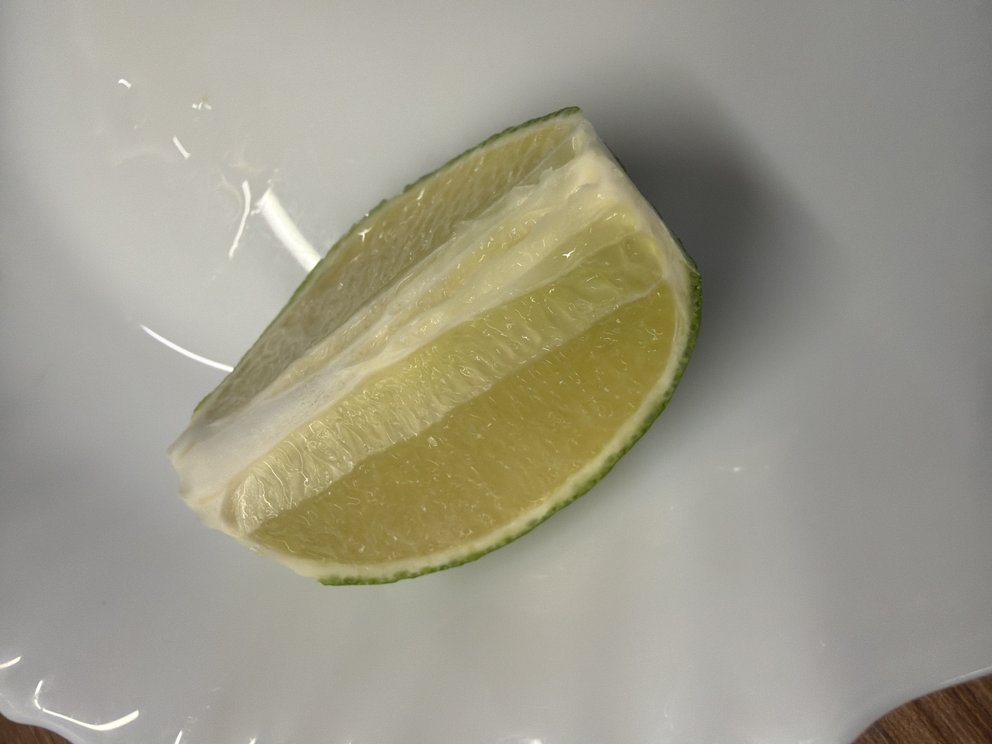 A wedge of lime sits on a white, textured plate. The lime is a vibrant green on the rind, transitioning to a juicy, yellow-green interior with visible segments and pulp. The plate has a slightly rippled edge. The background is a plain white surface with subtle reflections and some blurred areas, suggesting an indoor setting. There is a hint of a wooden surface at the bottom left of the image. No people or specific landmarks of Łódź, Poland are visible.