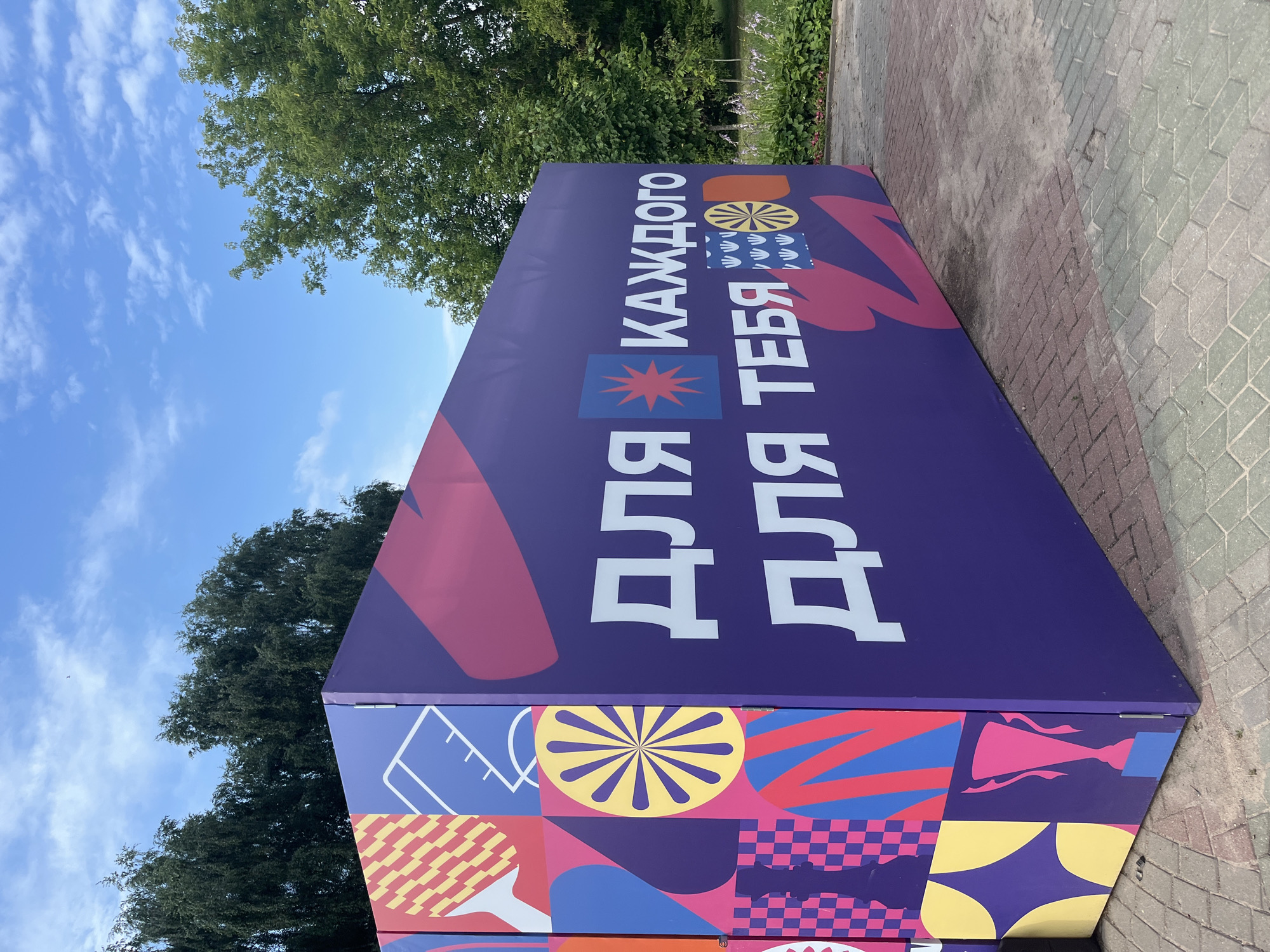 A large, brightly colored purple rectangular structure is situated outdoors on a paved area. The structure features a vibrant, geometric design with various shapes and patterns in shades of pink, yellow, blue, and red. Prominently displayed on one side of the structure in large white Cyrillic lettering are the phrases "ДЛЯ КАЖДОГО" (for everyone) and "ДЛЯ ТЕБЯ" (for you). To the left, the structure's side panel is covered with a collage of abstract graphics, including what appears to be a tennis racket, a chess piece, a geometric sun-like shape, and a trophy. Behind the structure, a line of tall, dark green trees forms a backdrop, with a bright blue sky dotted with white clouds visible above. In the distance, a patch of green foliage with some light-colored flowers is visible to the right of the structure. The ground beneath the structure is paved with interlocking bricks in varying shades of red, gray, and green. This structure is likely part of an outdoor exhibition or event.