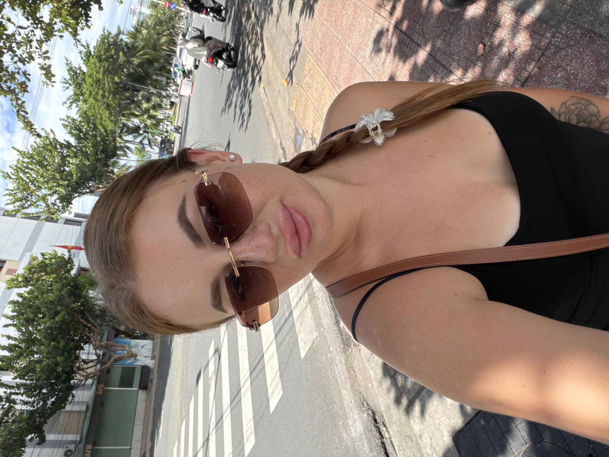The image is a selfie-style portrait of a young woman taken outdoors on a sunny day. The woman is positioned centrally, looking directly at the camera. She has light brown hair styled in a braid with a white flower-shaped clip, wears brown-tinted sunglasses with a gold frame, and a black top with a brown strap visible over her shoulder. A small pearl earring adorns her left ear, and a floral tattoo is visible on her right shoulder/upper arm. Her lips are slightly parted, and her expression is neutral.
The background reveals an urban street scene in Nha Trang Ward, Vietnam. Lush green trees, including palm trees, line the roadside, and several multi-story buildings with various facades, including one with "378" visible on it, stand along the street. A red and yellow flag, likely the Vietnamese flag, is also present near a building. The road features a painted white crosswalk. In the distance, motorcycles and cars can be seen, suggesting an active street with traffic. Strong sunlight casts distinct shadows on the pavement and sidewalk, indicating midday or early afternoon. The sky is bright and clear with some clouds.