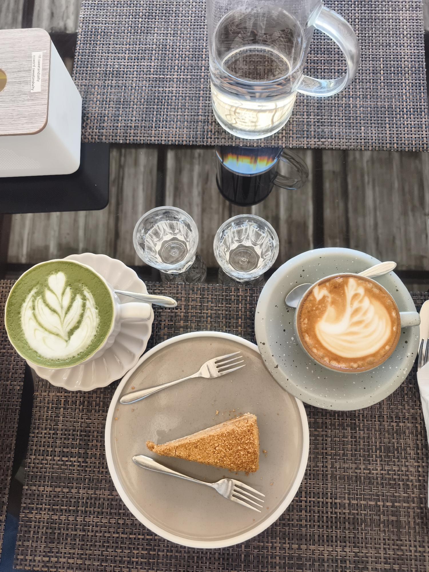 A high-angle, medium shot captures a cafe table set for a light meal. Two drinks, a matcha latte with intricate foam art and a latte with swan-shaped foam art, are presented in cups on their respective saucers. A slice of cake, coated in finely crushed nuts, sits on a gray plate with two forks. Two small glasses of water are also visible, alongside a larger pitcher of water and a tissue box. The table is covered with a dark woven placemat and has a dark wood grain surface visible beneath. The scene is set in Baan Mae Nam, Thailand.