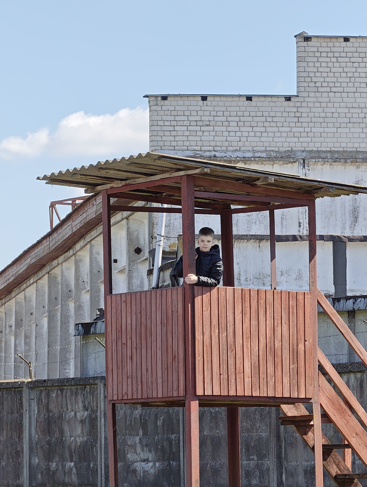 A young boy stands in a red wooden guard tower with a corrugated metal roof. The tower has wooden stairs leading up to the platform where the boy is standing. The boy is wearing a black jacket and appears to be looking directly at the camera. Behind the tower is a large concrete building with barred windows and a brick extension on top. A barbed wire fence runs along the base of the concrete building. The sky is blue with some clouds. The location is Hrodna, Belarus.