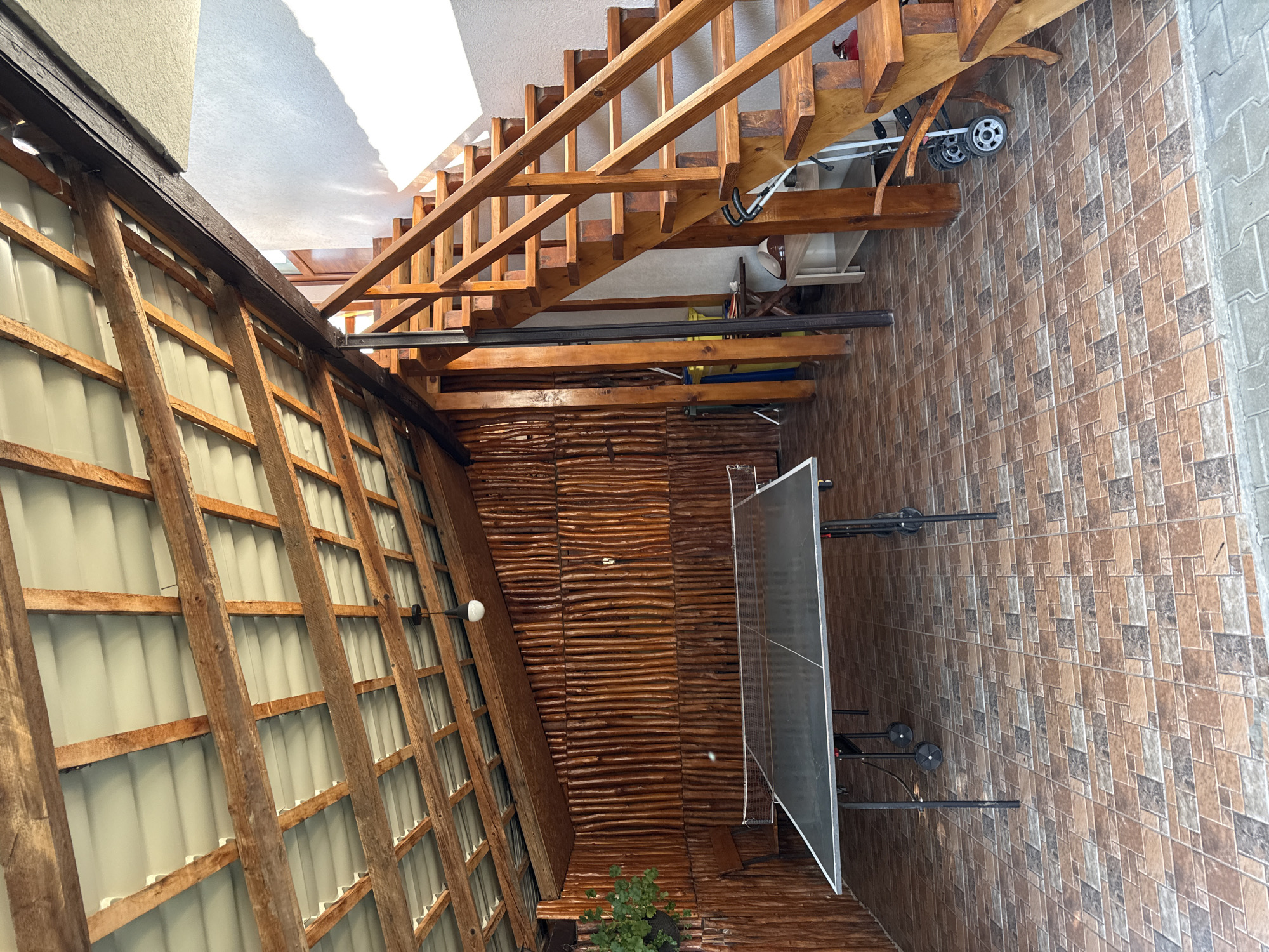 An outdoor covered patio area is shown, featuring a ping pong table in the foreground. To the right, a wooden staircase ascends. The walls are paneled with vertical wooden logs. The ceiling consists of exposed wooden beams supporting a corrugated metal roof. The floor is tiled in a mosaic pattern of brown and beige squares. A potted plant hangs from the upper left. The setting appears to be within a property in Cârțișoara, Romania.