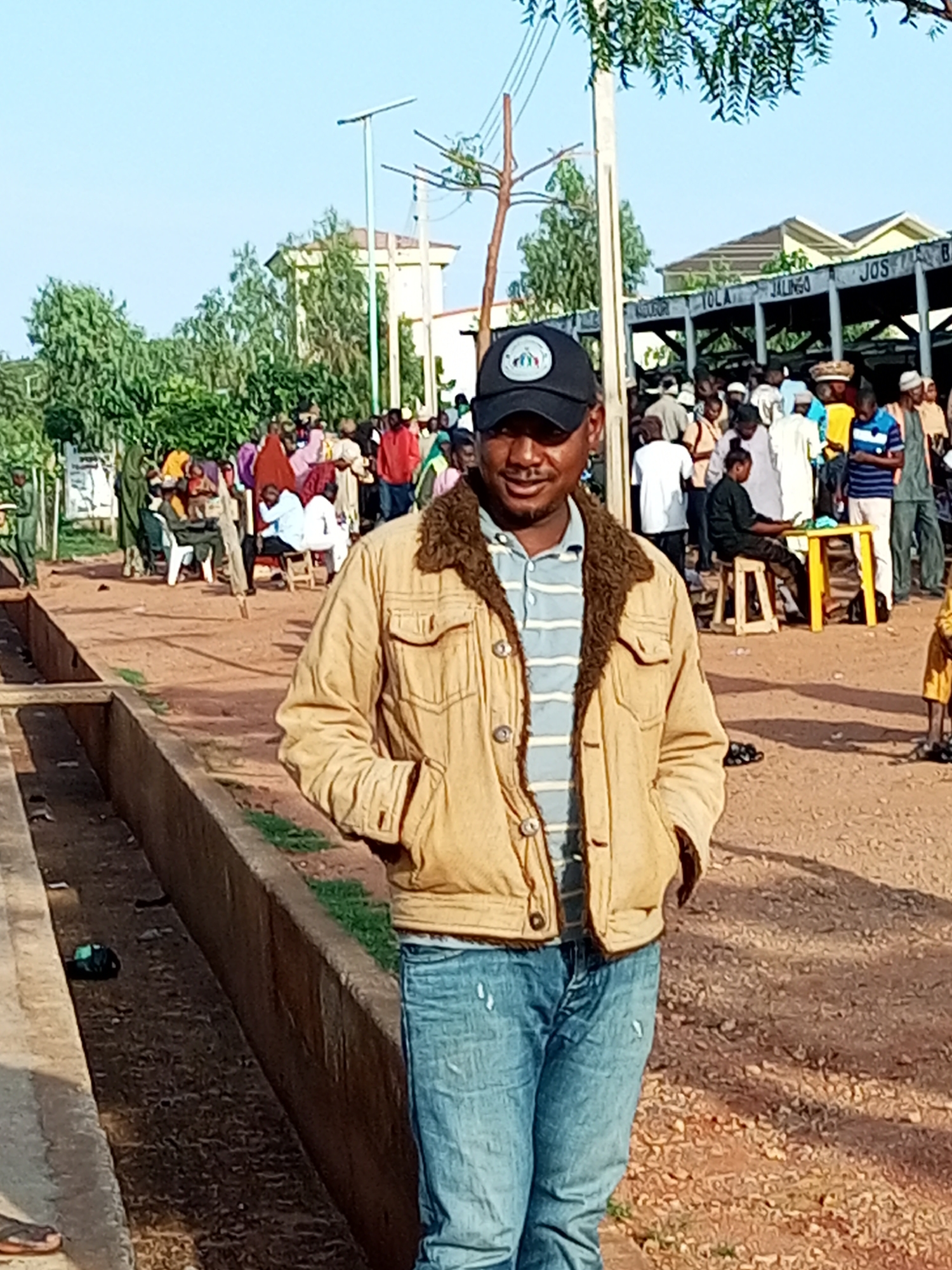 The image shows a man standing in the foreground, wearing a tan jacket with a fur-lined collar, a striped shirt, and blue jeans. He is also wearing a black baseball cap. Behind him, a crowd of people are gathered. In the background, there is a building with signs that appear to list city names, including "YOLA," "JALINGO," "JOS," and "BA." This suggests the location may be a transportation hub or a place where people are gathering for travel. The environment appears to be outdoors, with dusty ground and some vegetation visible. The overall scene suggests a busy public area. The presence of the city names on the building strongly indicates the location is the Gombe Central Motor Park in Gombe, Nigeria.