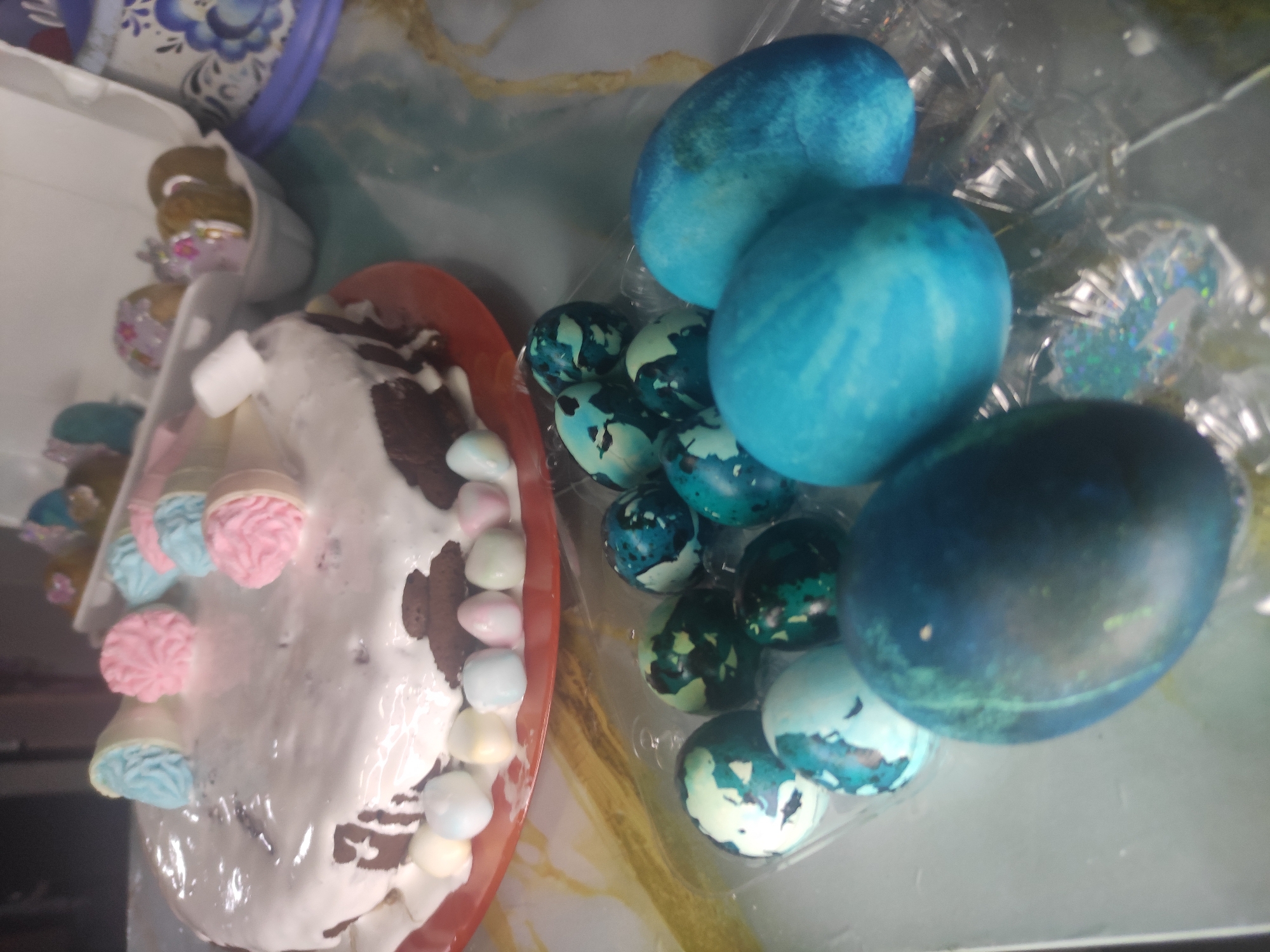 The image shows an Easter celebration scene. There is a decorated Easter cake covered in white frosting, decorated with meringue and marshmallows and with brown cake visible underneath the frosting. Next to the cake are decorated Easter eggs, some dyed shades of blue. Others are decorated with teal, green and white patterns. The scene is in Chaikovsky, Russia.