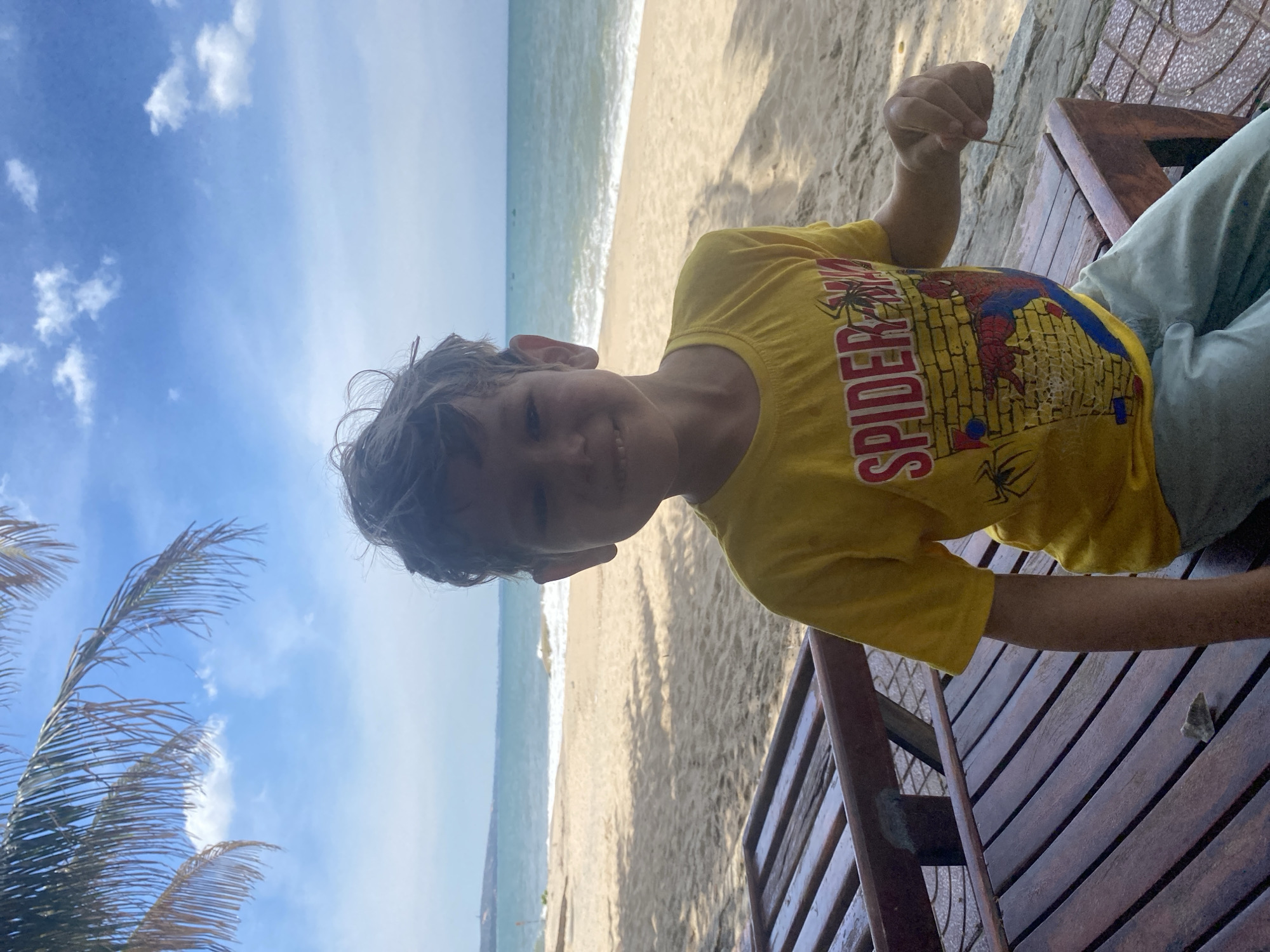 A young boy with tousled brown hair smiles at the camera while sitting on a wooden bench on a sandy beach. He is wearing a yellow t-shirt with a Spider-Man graphic and light blue pants. Behind him, the ocean stretches to the horizon under a blue sky with scattered clouds. Palm fronds hang overhead, casting shadows on the scene. The beach is mostly empty, with a few small boats visible in the distance on the water. The overall impression is one of a relaxed, sunny day at the coast. The location appears to be Phu Thuy Ward, Vietnam, known for its beautiful beaches and coastal scenery.