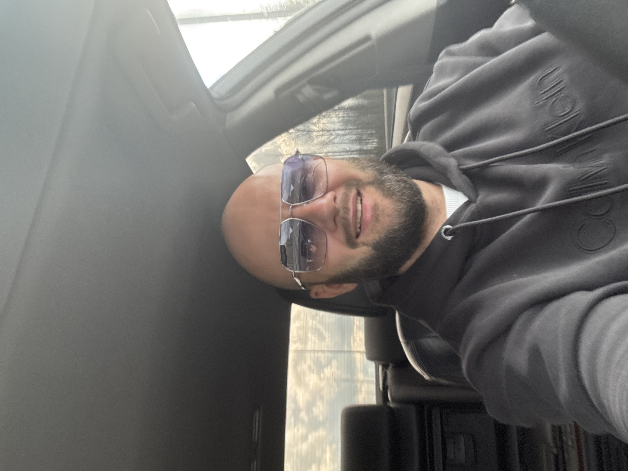 A person is sitting inside a vehicle, wearing a dark grey hoodie with the brand "Calvin Klein" embroidered on the front. The individual is bald and has a beard, and is wearing sunglasses. The interior of the car is visible, including the door panel and parts of the dashboard and seats. The vehicle's window is open, revealing a view of the outside, where the scene is unidentifiable and the city and location are unknown.