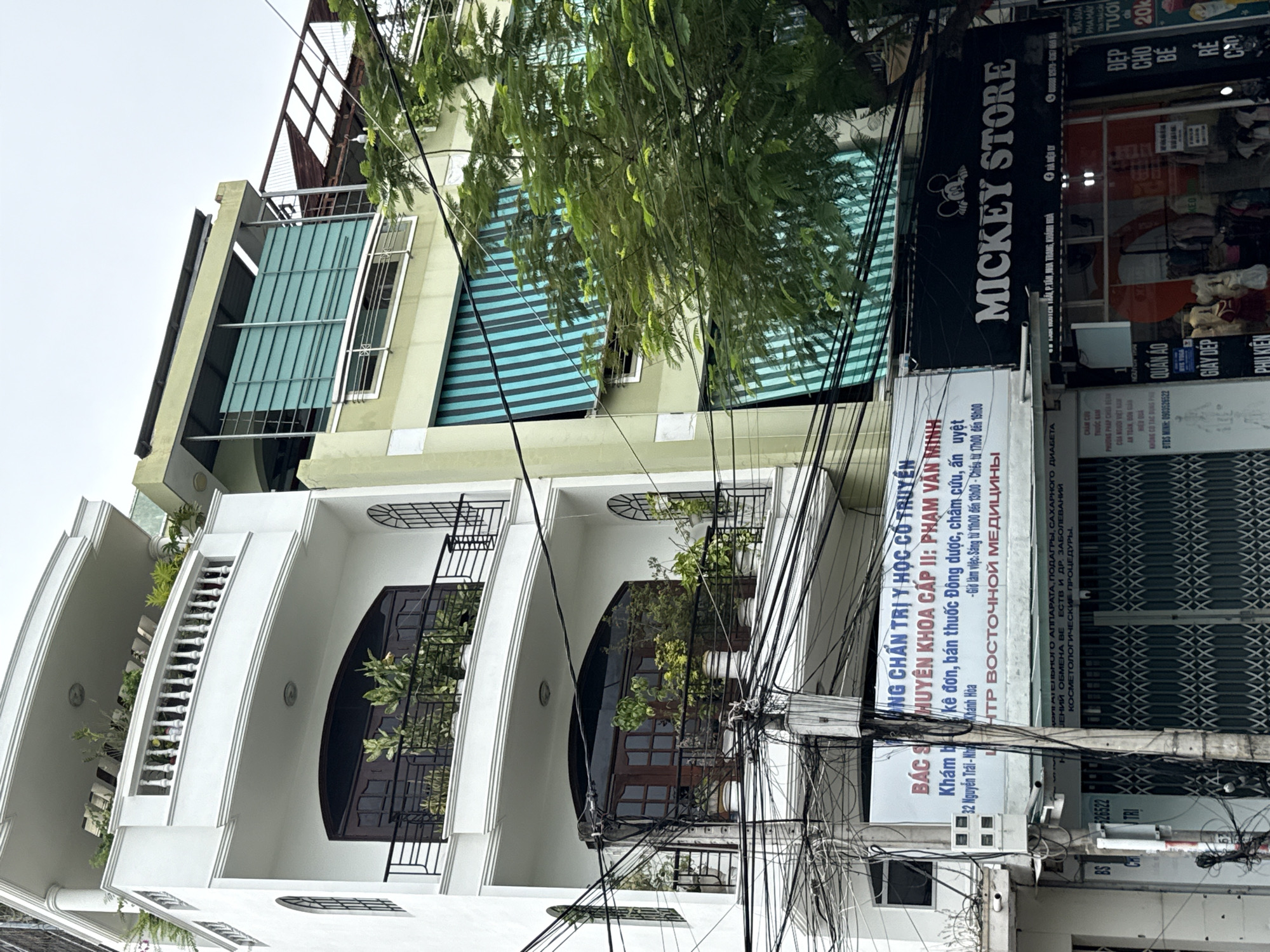 The image shows a street scene in Nha Trang, Vietnam. In the foreground, a white building with arched windows and balconies with potted plants is visible. A complex tangle of electrical wires hangs in front of the building. To the right of the white building, a light green building with blue and white striped awnings is partially visible. A large tree with green foliage obscures a portion of the buildings. Several signs are mounted on the lower part of the buildings. One prominent sign reads "MICKEY STORE" in white letters on a black background, featuring an image of Mickey Mouse. Below this, a store interior with mannequins displaying clothing is visible. Other signs display text in Vietnamese and Russian. The sky is overcast.