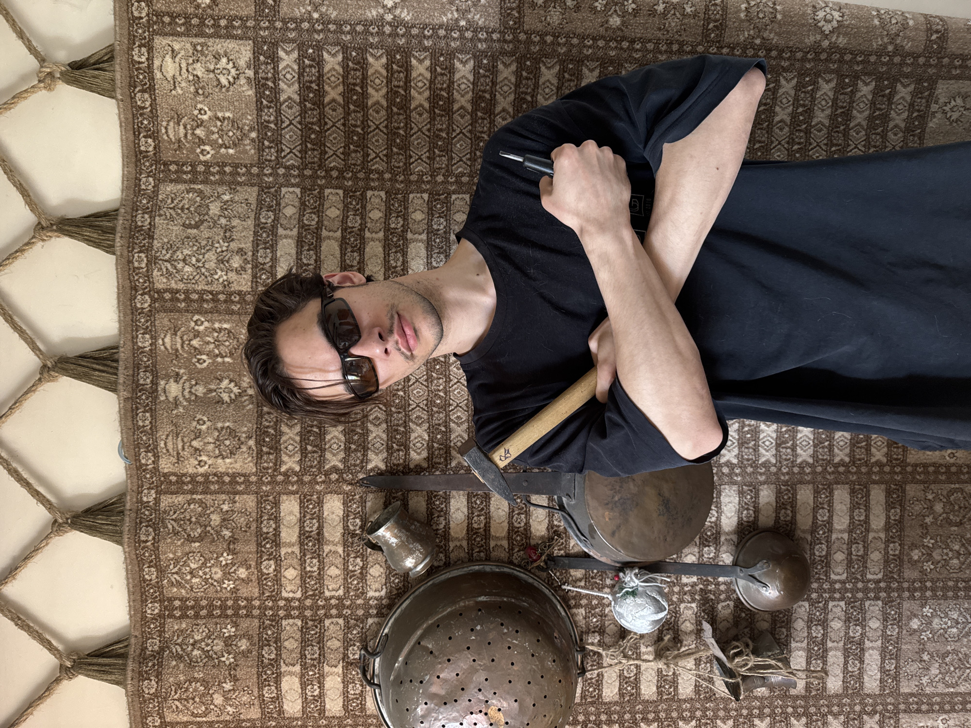 A young man with dark hair, a light mustache, and dark sunglasses lies on his back on a patterned brown and beige rug. He is wearing a black t-shirt with a faint, unreadable graphic on the left side. His arms are crossed over his chest, with his right hand holding a black screwdriver or small tool. He looks directly at the camera with a neutral expression.
Arranged around his lower body and on the rug are several antique-looking metal objects. These include a large, round, hammered copper colander with handles, a small ornate silver or pewter pitcher, and a dark, round metal pot. A long, narrow metal blade, possibly part of an old tool, rests alongside a wooden handle resembling a pickaxe, which has a carved symbol like a stylized 'E' or '3'. Other items visible are a long-handled metal ladle, a small spherical object wrapped in white/silver material with string, and various other small metal pieces and twine.
The rug features traditional patterns and has tassels along its left edge, next to a light-colored wall. The scene is brightly lit, suggesting daytime, and gives a rustic or antique aesthetic. The location context is Sevastopol, Ukraine.