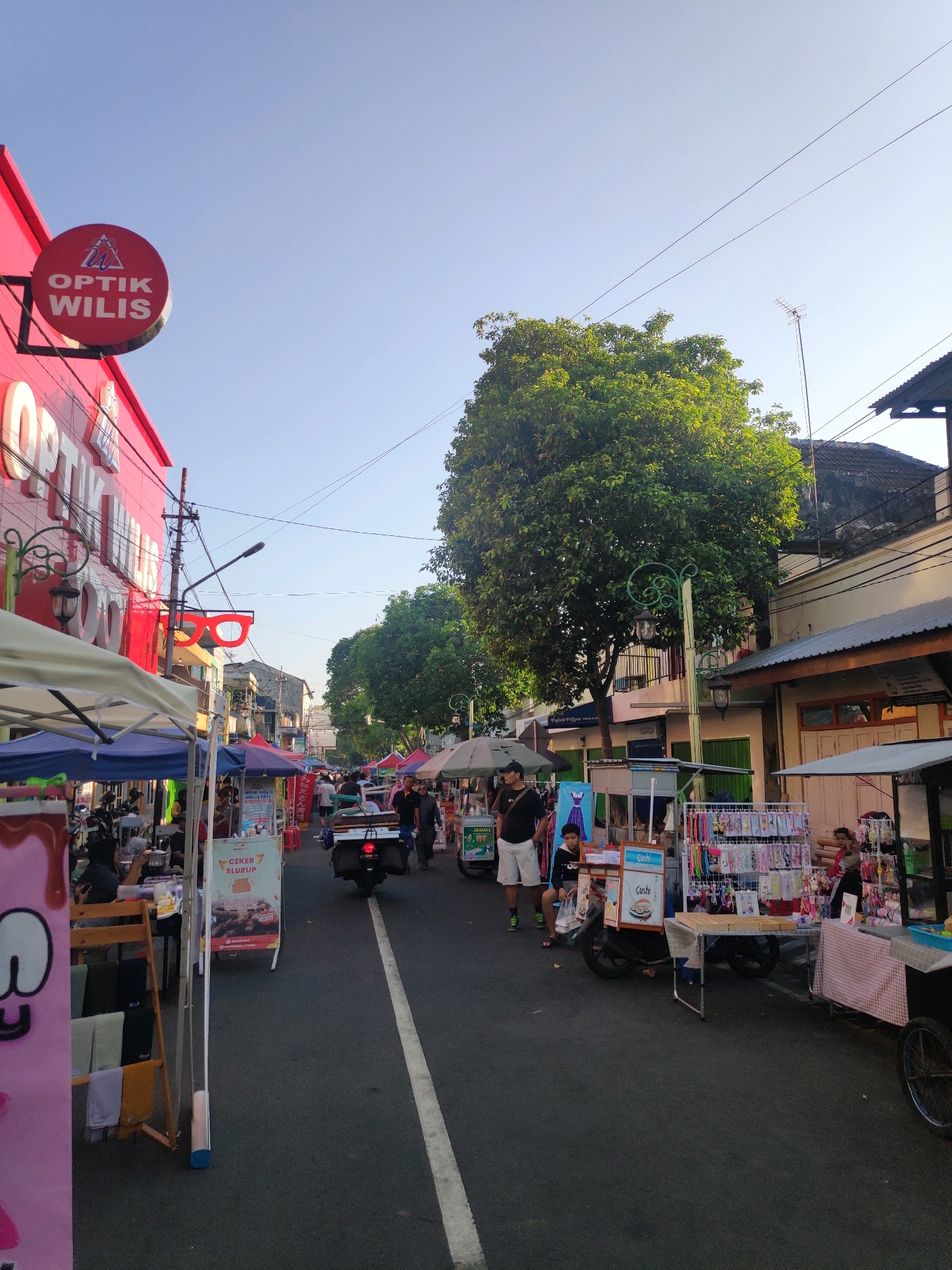 The image shows a street scene in Kebonagung, Indonesia, likely a market or a street fair. The foreground features several market stalls, some with awnings and displays of goods such as clothing and accessories. A pink tarp with a white graphic is visible on the left. In the middle ground, a street runs through the scene, with people walking and a motorcycle with a passenger carrying items. One of the stalls has a sign advertising "Ceker Slurrp". On the left side of the street, a building with a red facade displays the signage "OPTK WILIS" and a circular sign with glasses and the text "OPTK WILIS". A decorative street lamp in the shape of large red glasses is also visible. The background shows more buildings, trees, and power lines under a clear blue sky. The lighting suggests it is either early morning or late afternoon.