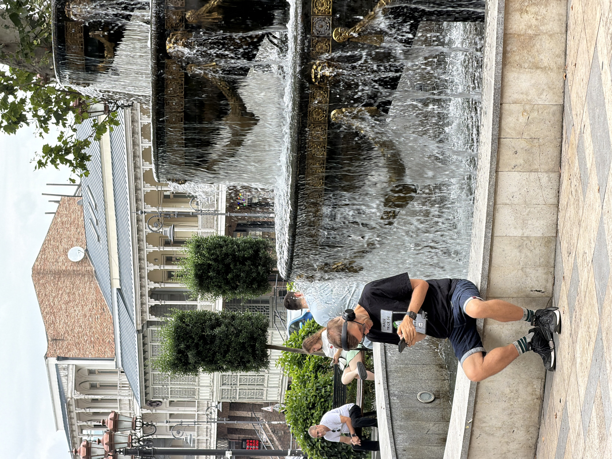 A man with headphones on is sitting on a fountain ledge, looking at his phone. He is wearing a black t-shirt, denim shorts, striped socks, and black sneakers. To his left, a woman is sitting on a bench, also looking at her phone. Further left, a man in a white t-shirt and black pants is sitting on the same bench. Behind them, a large multi-tiered fountain with golden accents is cascading water. In the background, there are traditional Tbilisi-style buildings with ornate balconies and a brick facade. The sky is overcast.