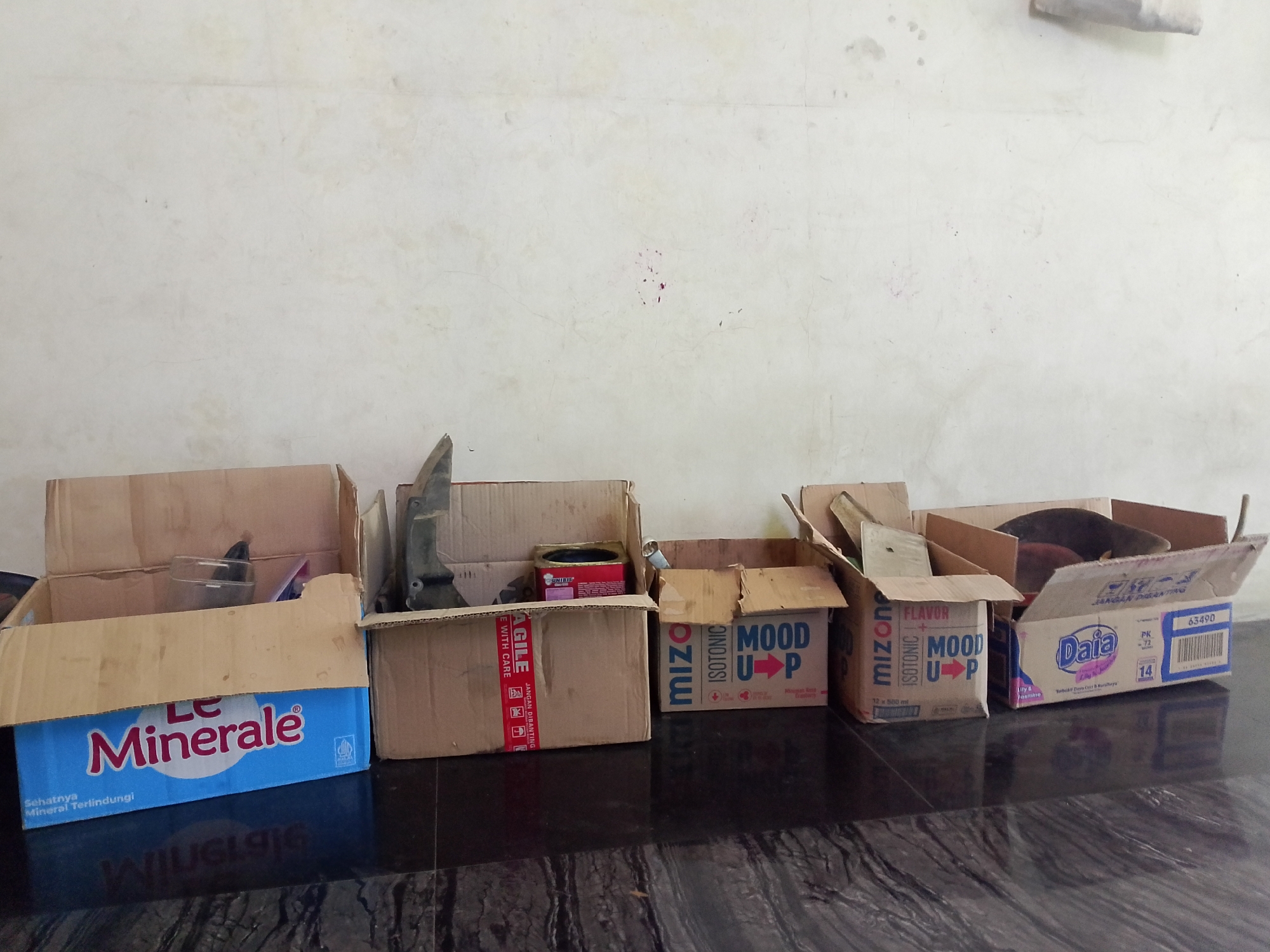 In Situwangi, Indonesia, a line of cardboard boxes is placed against a light-colored wall. The boxes are of varying sizes and contain various items, including a glass, a red tin, and what appears to be a black animal, possibly a cat, nestled within one of the boxes. One box displays the "Le Minerale" brand logo. The floor is a dark, reflective surface, possibly polished stone or tile. The scene suggests a storage area or a collection of unpacked items.