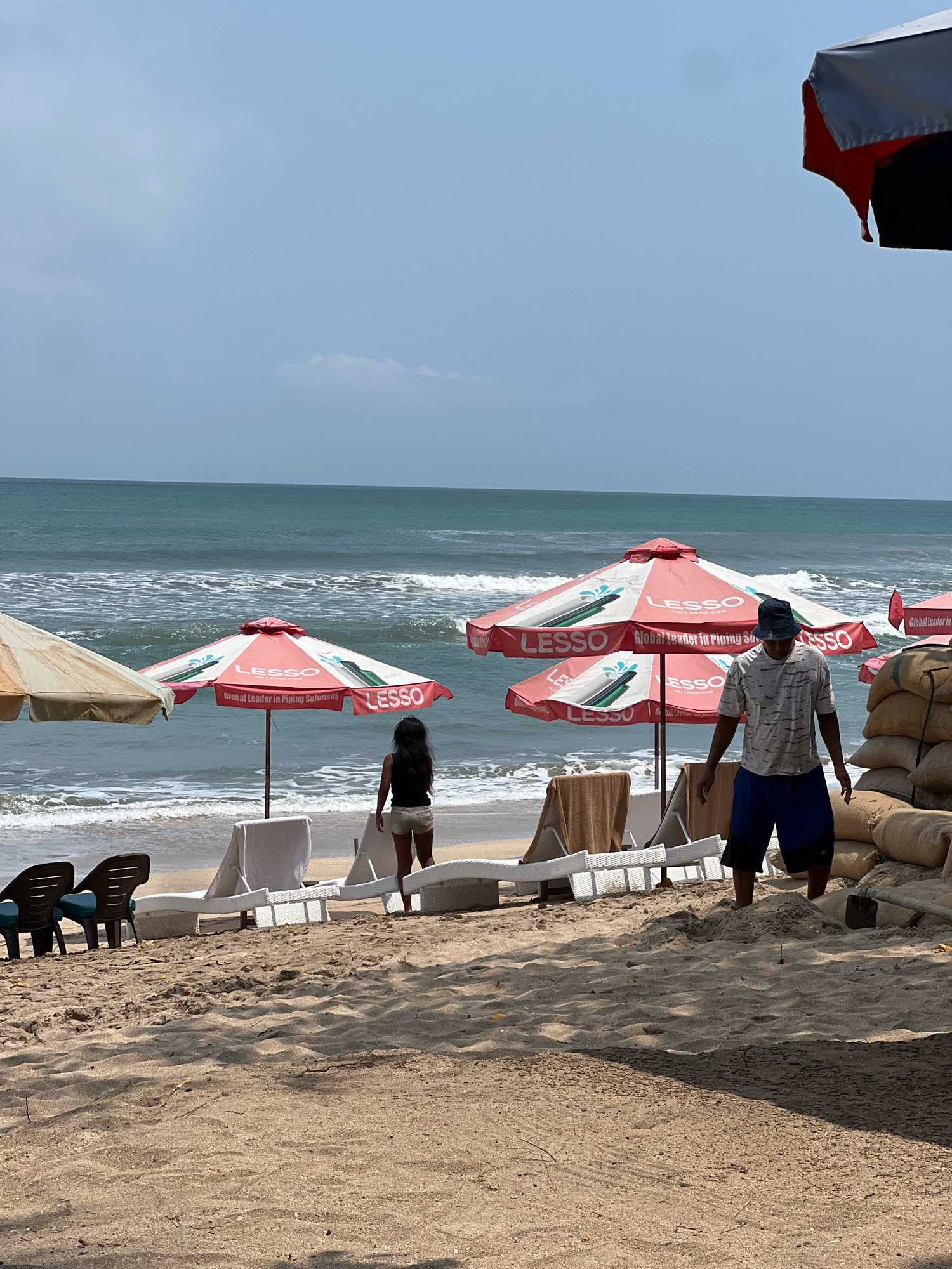 The image depicts a sunny day at Kuta Beach in Bali, Indonesia. The foreground is dominated by sand, with some footprints and natural debris visible. A man wearing a dark bucket hat, a patterned t-shirt, and blue shorts is walking on the sand. To his left, there are stacks of burlap sacks, some with a shovel propped against them.
In the mid-ground, a beach scene unfolds with several beach umbrellas, two of which are prominent and feature red and white branding with the text "LESSO Global Leader in Piping Solutions." White lounge chairs are arranged on the sand, some covered with towels. A woman with dark hair, wearing a black tank top and light-colored shorts, is standing near one of the umbrellas, facing the ocean.
The ocean itself is visible in the background, with gentle waves breaking on the shore. The water appears blue-green. The sky is clear with some scattered clouds. To the far left, another umbrella, this one beige, is partially visible. The scene suggests a typical day at a popular tourist beach.