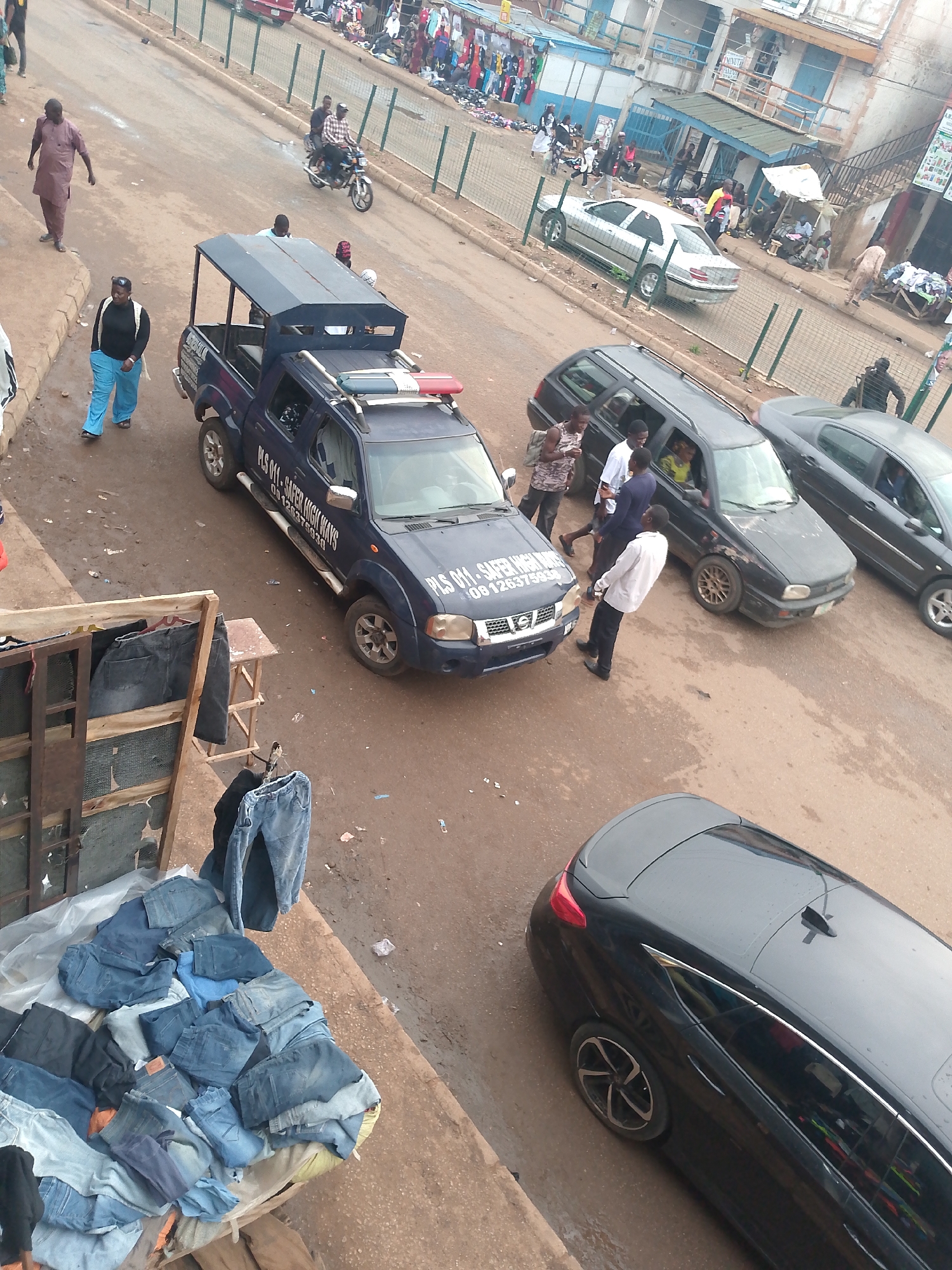 The image, taken from an elevated perspective in Bukuru, Nigeria, depicts a busy street scene. A blue police pickup truck with a black roof is parked, with two men standing beside it. Three other cars are parked nearby: a dark station wagon, a dark sedan, and a black sedan. Several people are walking on the street and sidewalk, including a man in a brown outfit and a woman in a blue tracksuit. A motorcycle is being driven on the road in the background. Merchandise, likely clothing, is displayed on a stand in the foreground. Buildings and shops line the street, and a fence separates the road from a pedestrian area in the background.