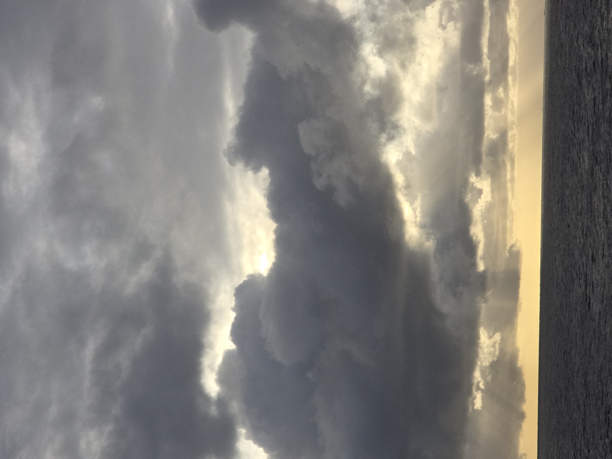 The image depicts a sky filled with predominantly gray clouds, with the sun partially obscured, creating a play of light and shadow. To the right, a body of water is visible. The scene appears to be a waterfront view at either sunrise or sunset. The location is unknown.