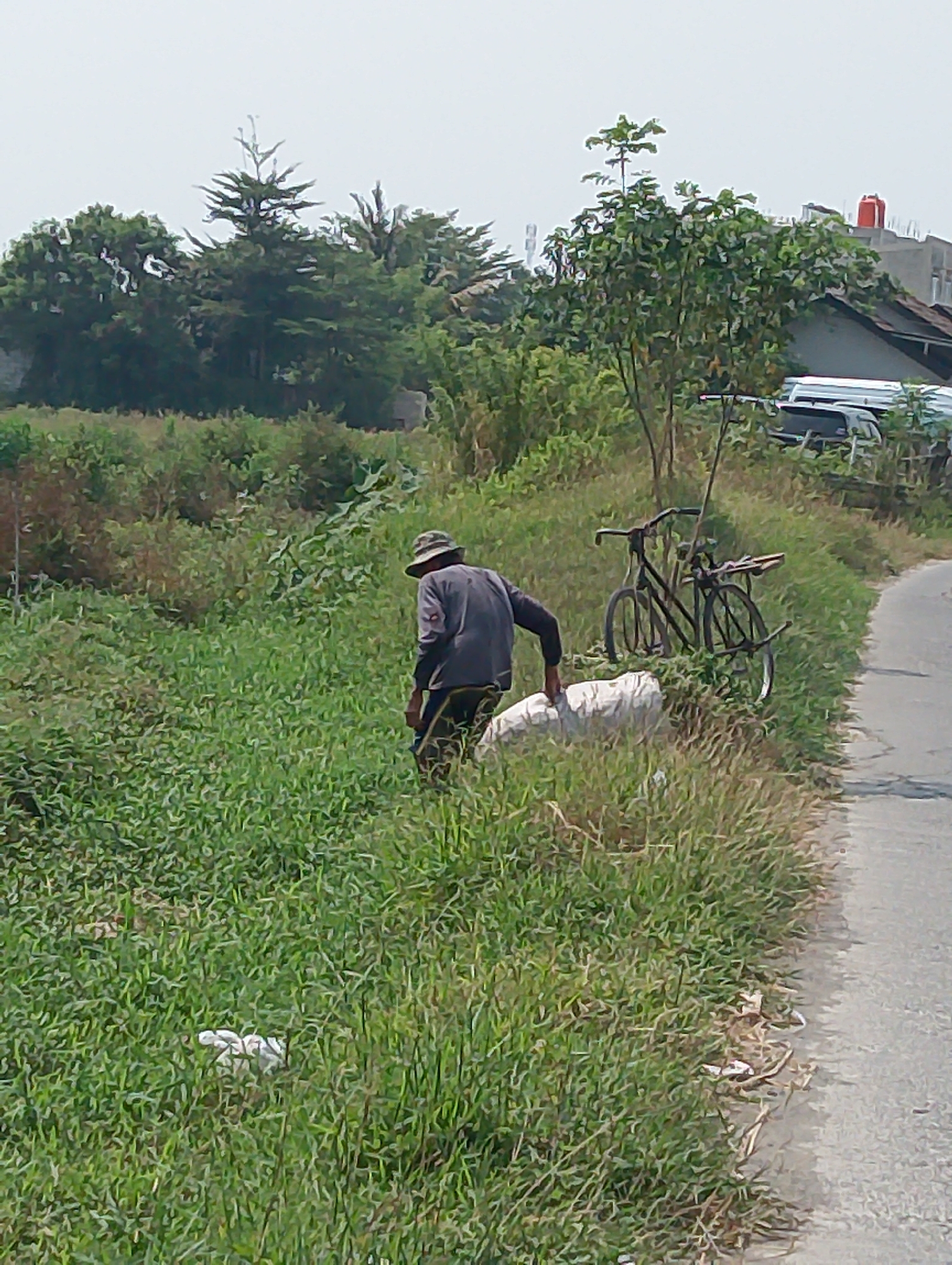 A man wearing a hat and a dark long-sleeved shirt is carrying a large white sack. He is standing in a grassy area next to a road. A bicycle is leaning against some bushes behind him. In the background, there are trees, houses, and a red water tank. The sky is overcast. The scene appears to be in a rural or semi-rural area of Karawang, Indonesia.