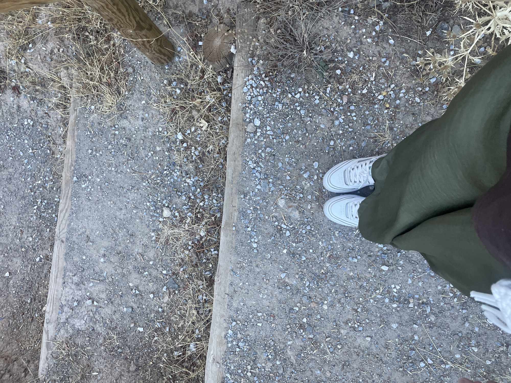 The image shows a person's feet wearing white sneakers and a green skirt standing on a gravel path with wooden steps. The path appears to be outdoors, possibly in a natural setting, with dry grass and vegetation around. In the background, a wooden post is visible, suggesting part of a railing or fence. The lighting indicates it is daytime. The location is identified as the city of Víznar, Spain.