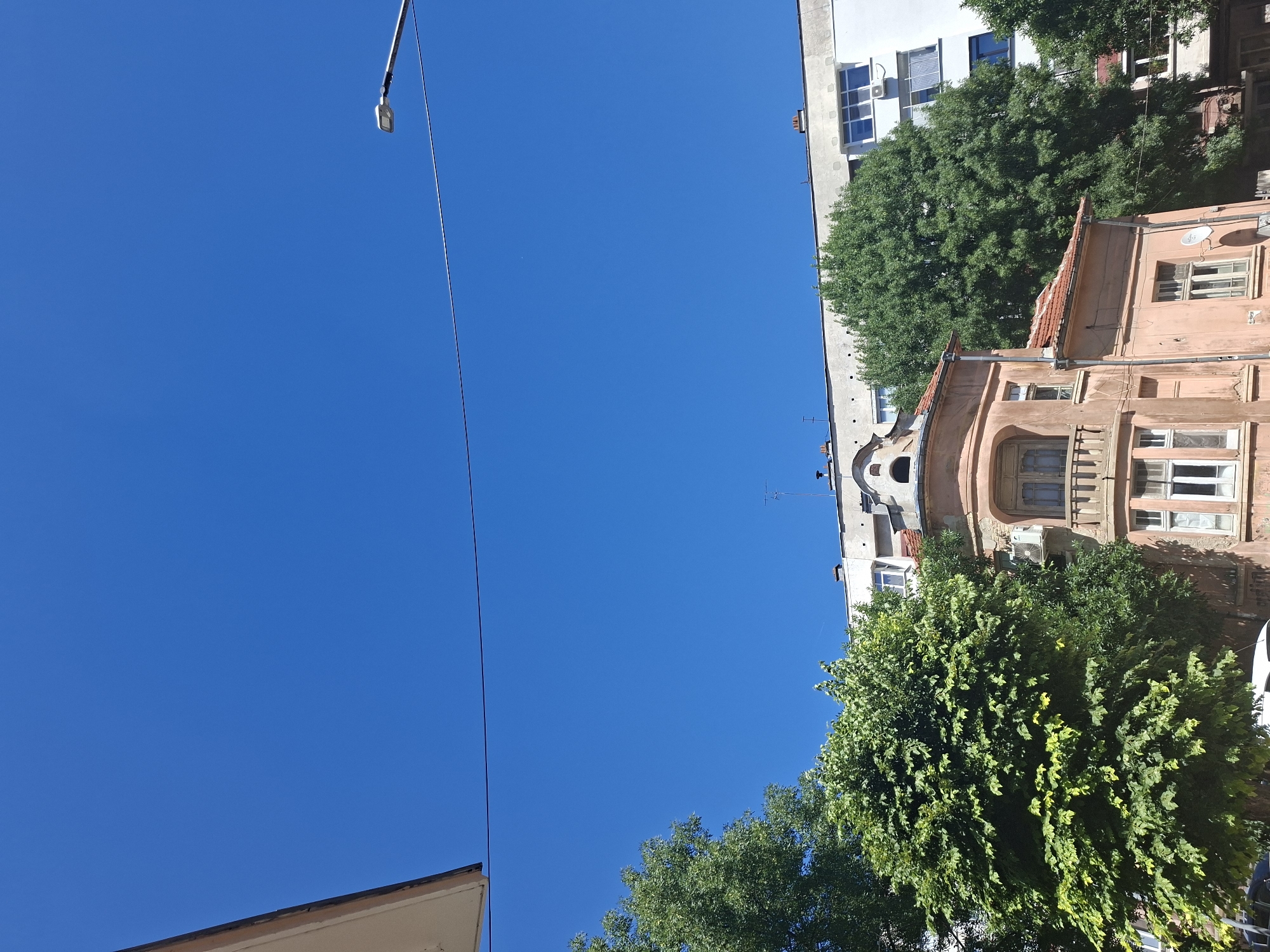 The image depicts a clear blue sky with a few trees and buildings in the foreground. The buildings appear to be a mix of older and newer styles. One older, pale pink building with ornate architectural details stands out, possibly an older residential building or a historic site. Behind it, a larger, modern white building with visible windows and air conditioning units is visible. The trees are lush and green, suggesting a warm season. A street lamp is visible on the right side of the image. The setting is likely an urban environment. The image can be located in Pleven, Bulgaria, given the context.
