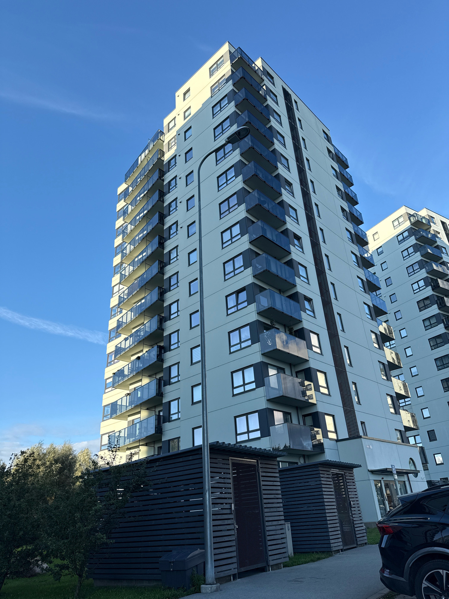 This is a low-angle, full-shot photograph of modern apartment buildings in Tallinn, Estonia, under a clear blue sky with wispy clouds. The main building, a tall, light-colored structure, dominates the left side of the frame, featuring multiple floors with dark-paneled balconies and numerous rectangular windows. To the right, a similar, slightly shorter building is partially visible. In the foreground, there are dark wooden structures that appear to be storage units or sheds, and a portion of a black car is parked on the right. Green trees and grass are visible on the left. The scene suggests a residential area with contemporary architecture.