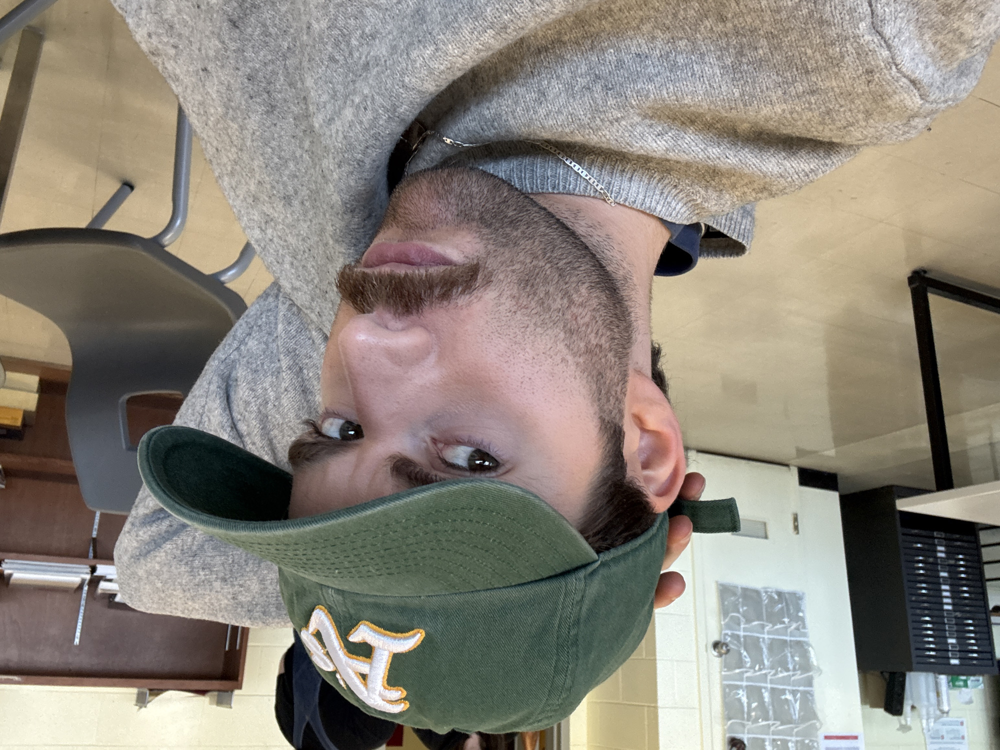 A young man is captured in a close-up, headshot-style image, looking directly into the camera. He has facial hair, including a mustache and beard, and is wearing a light grey knitted sweater. Around his neck, a thin silver chain is visible. He dons a dark green baseball cap, prominently featuring the "A's" logo in white with a yellow outline on the front. His eyes are dark and focused, conveying a direct and somewhat intense gaze.
The setting appears to be indoors, possibly a classroom or an institutional building. In the background to the man's left, parts of a dark grey chair with metal legs and a wooden surface with stacked papers or books are visible. To his right, a white door with a small window and handle is present, along with a clear plastic shoe organizer with multiple pockets hanging on it. Further right, a dark rectangular object with horizontal slats, possibly a storage unit or equipment, is partially visible. The floor is a light, neutral color. The overall lighting suggests an indoor environment without specific time-of-day indicators. The image's context is Montreal, Canada.