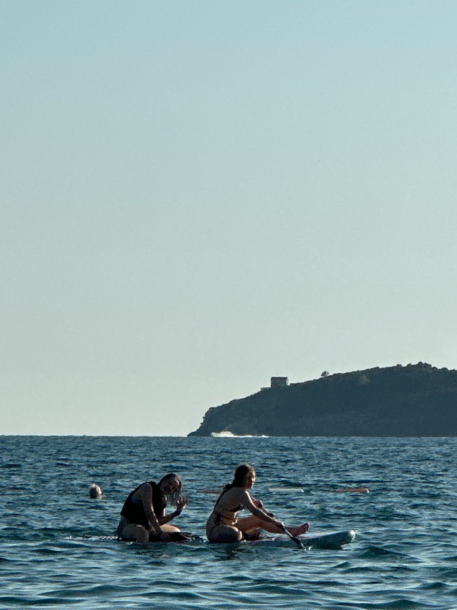 Two individuals are seated on paddleboards in the sea, with a dark, tree-covered island in the background. The island features a small, light-colored structure on its highest point, possibly a tower or old building. The water is choppy and a light-colored buoy is visible in the water between the two paddleboards. The sky is a clear, pale blue, suggesting a sunny day. The scene appears to be near the city of Boreti, Montenegro, given the coastal topography and the presence of such islands.