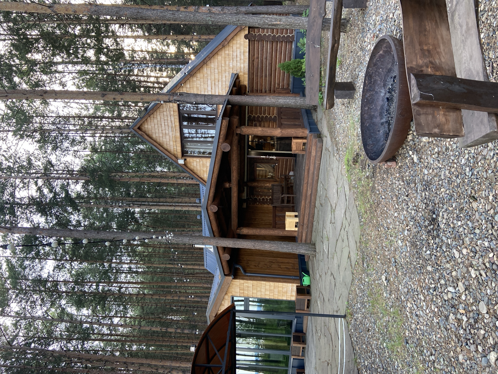 The image depicts a rustic wooden house surrounded by a dense pine forest. The house has a traditional log cabin style with a prominent porch and a steeply pitched roof. The exterior is clad in natural wood shingles and logs. In front of the house, there is a paved patio area with stone tiles. A fire pit made of dark metal sits in the foreground, surrounded by gravel and several wooden benches. String lights are visible in the trees, suggesting an outdoor seating or entertainment area. The environment is natural and serene, typical of a forest setting. The location is specified as Забегаево, Russia. There are no people visible in the image.
