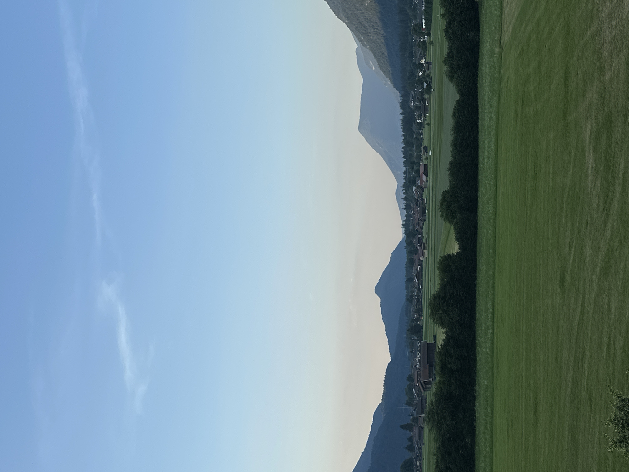 This is a vertical, wide-angle shot of a scenic landscape in Garmisch-Partenkirchen, Germany. The image is divided into two primary sections: the sky and the ground. The sky dominates the left side of the frame, displaying a gradient of pale blue, lighter at the horizon and deepening towards the top. A few wispy, white clouds are scattered across the blue expanse.
The right side of the image reveals a mountain range, its peaks silhouetted against the sky. The mountains appear to be covered in dark vegetation. Nestled at the base of the mountains is a village, characterized by a dense line of houses with dark roofs. A row of lush, dark green trees separates the village from a vast, vibrant green field that stretches across the rightmost portion of the frame. The field appears to be a mowed lawn or pasture, with subtle variations in its texture and shading.
The overall impression is one of a tranquil, idyllic rural setting. The lighting suggests it is either early morning or late afternoon, with the sun casting a soft glow on the landscape. The clear sky and verdant fields evoke a sense of peace and natural beauty. There is no visible text or identifiable activity beyond the static presence of the village and the natural elements. The image captures a classic alpine vista, showcasing the harmonious coexistence of human settlement and natural grandeur.
