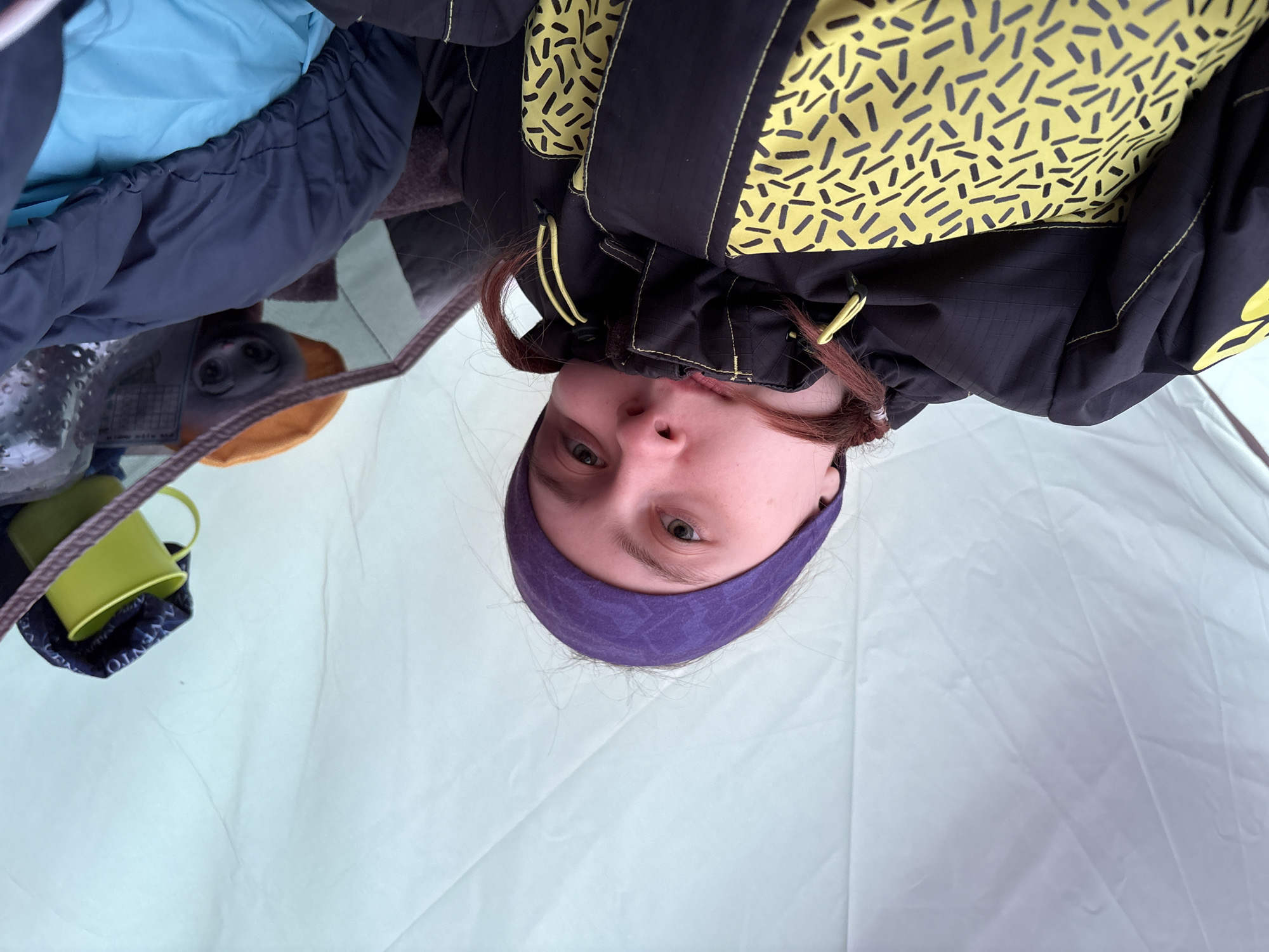 An upside-down close-up view captures a person looking directly into the camera from inside what appears to be a tent. The individual, likely female, wears a purple headband and is dressed in warm outdoor attire, including a black jacket featuring a striking yellow and black abstract pattern resembling sprinkles or dashes, layered over a dark blue puffy jacket. Their expression is neutral to slightly curious.
In the background, visible to the person's upper left, are various items indicating a camping or outdoor setting. These include a light blue fabric (possibly part of a sleeping bag or inner tent lining), a transparent plastic bottle, and a bright green metal cup. A dark blue fabric accessory with the white text "VENTO" printed on it is also visible near the cup. The dominant background is the light, somewhat wrinkled fabric of the tent's interior. A thin rope or strap crosses the top left corner of the frame. The unusual perspective suggests the photo was taken from a very low angle or with the camera inverted. The scene takes place in Lesnoye, Russia.