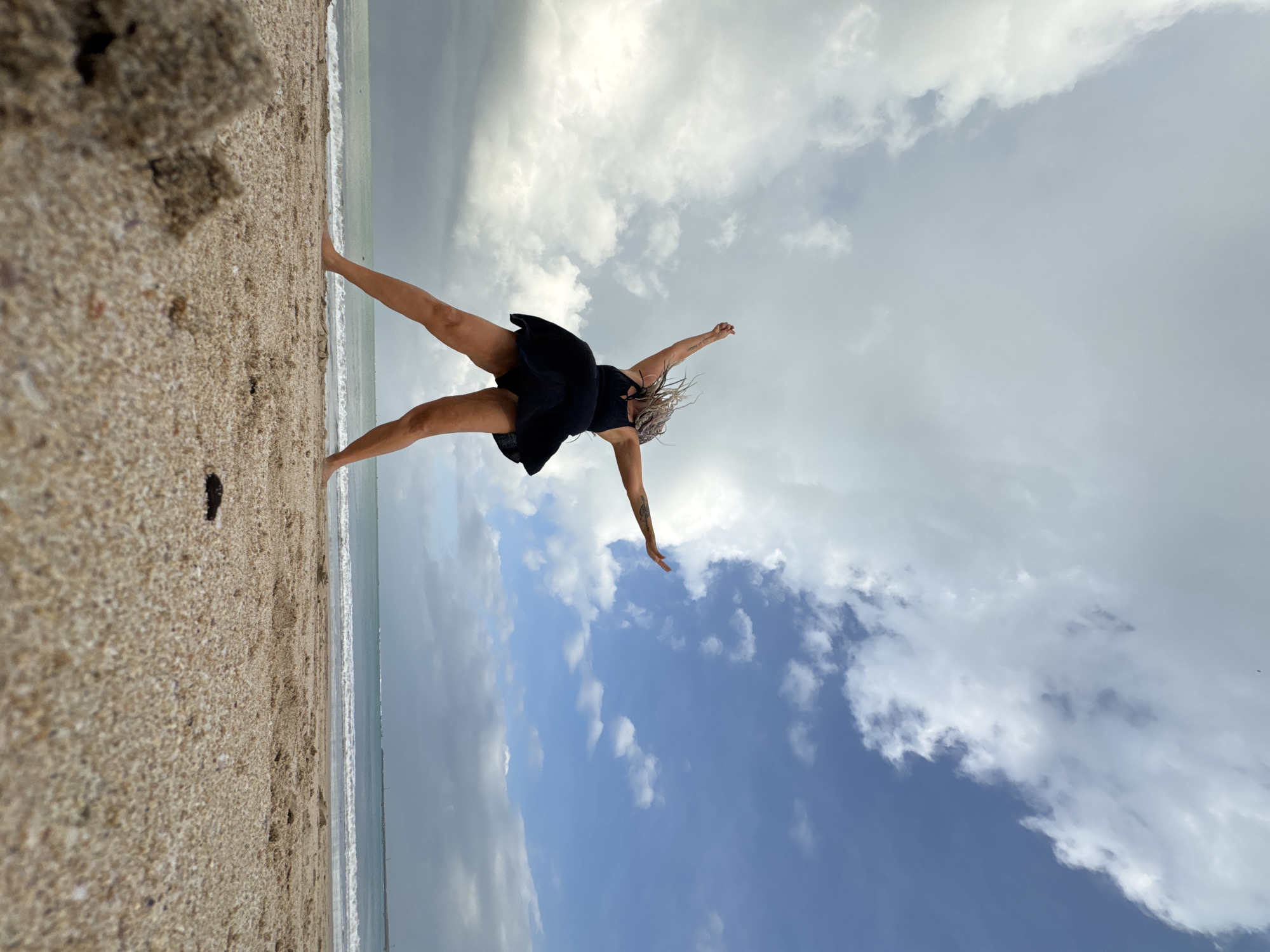 An individual is captured mid-jump on a sandy beach, their black dress billowing around them, arms outstretched against a dramatic sky of clouds and blue patches. The ocean is visible in the background with gentle waves. The low-angle perspective emphasizes the subject against the expansive sky, with sand in the foreground.