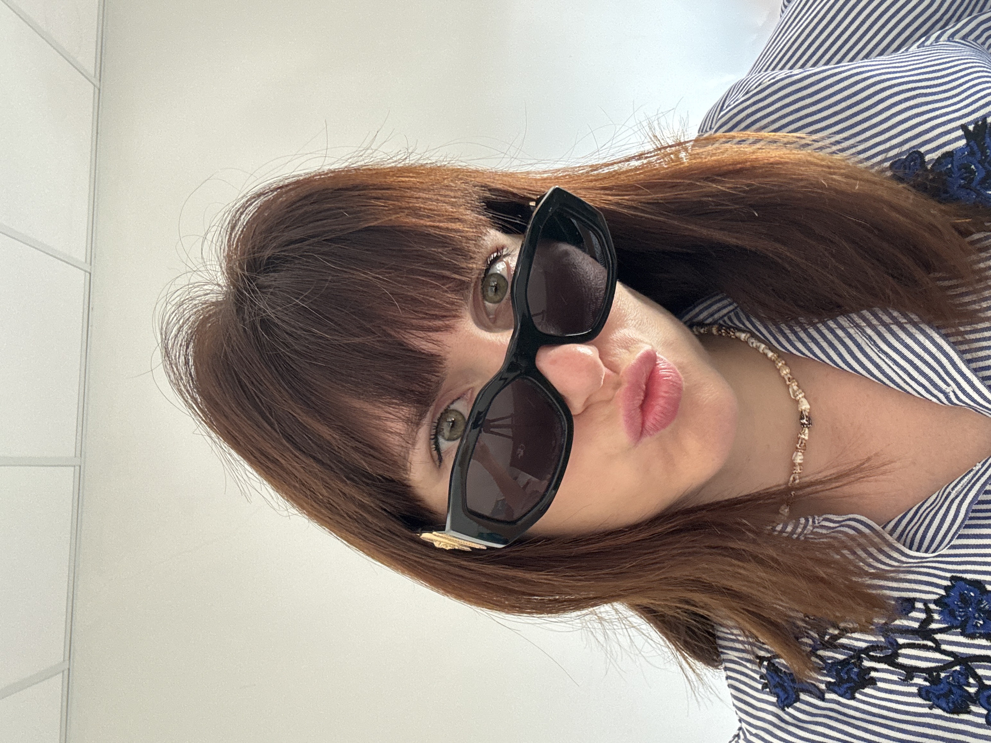The media file shows a close-up, waist-up portrait of a woman with auburn hair and bangs, wearing large, black-framed sunglasses and a blue and white striped shirt with floral embroidery. Her lips are slightly pursed, and she is wearing a beaded necklace. The lighting is bright, suggesting daylight, and the background is a plain, light-colored wall. The overall impression is that of a casual, personal snapshot, possibly taken indoors or in a well-lit outdoor space. There are no visible text elements in the image. The setting appears to be domestic or a casual public space. The woman's pose and expression suggest a playful or confident demeanor.