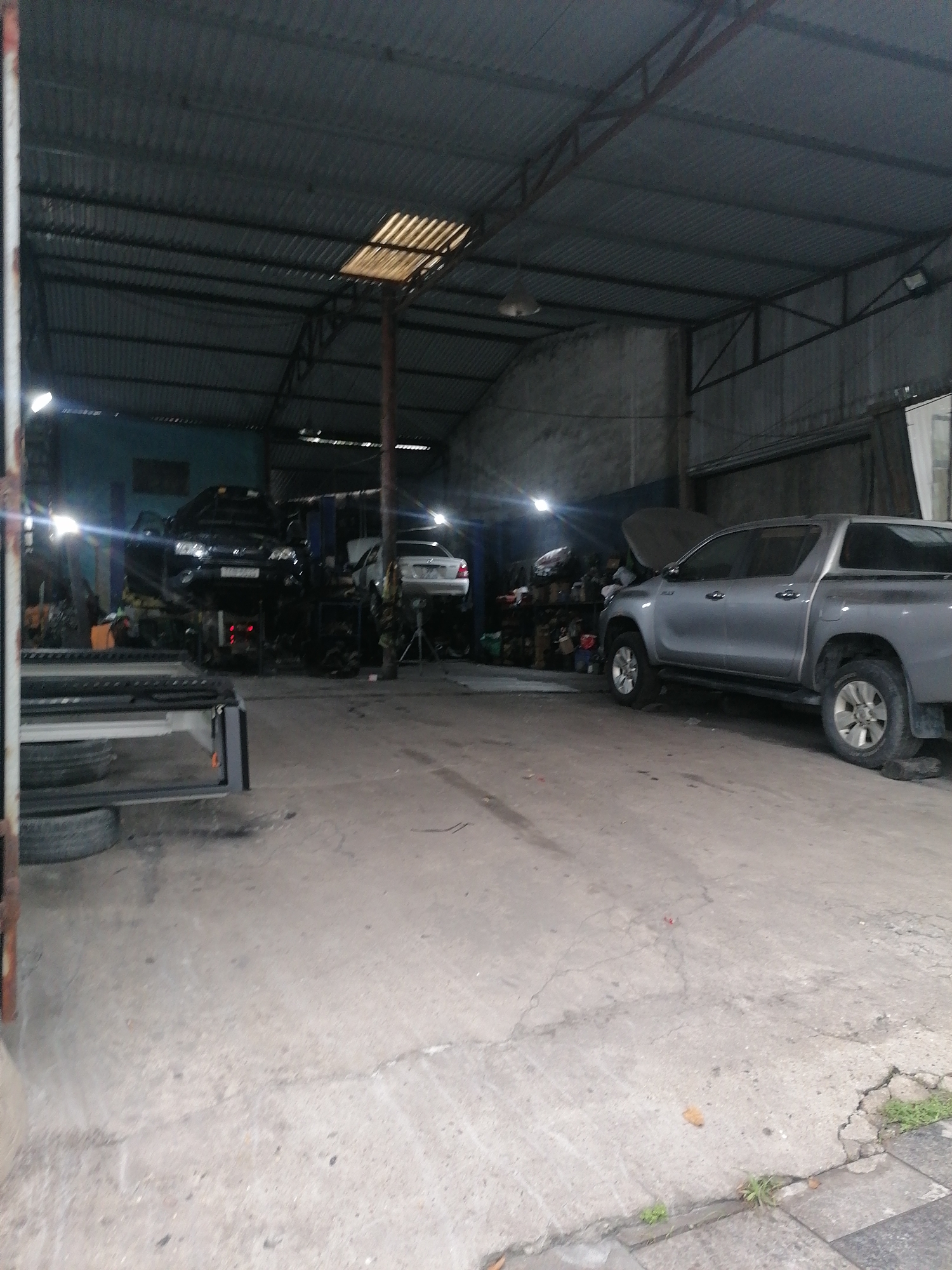 The image captures the interior of an auto repair garage, likely during the evening or night given the prominent use of artificial lighting. The scene features multiple vehicles in various stages of maintenance. On the left, a dark SUV or sedan is elevated on a hydraulic lift, with its hood open. Behind it, a white sedan is also seen raised, possibly on another lift or jack stands.
To the right, a silver pickup truck, similar to a Toyota Hilux, is parked with its hood open, indicating ongoing work. The garage has a robust, industrial structure featuring a corrugated metal roof supported by metal trusses and walls made of similar material. Bright artificial lights are visible throughout the space, creating some lens flares. The concrete floor appears worn, with visible cracks and some debris.
No individuals are present in the image. Various auto parts and equipment can be seen on shelves and scattered around the work areas. A part of what appears to be a vehicle chassis or trailer bed with a tire rests in the immediate foreground on the left. The overall impression is that of an active, functional vehicle repair establishment. No visible text is discernible in the image. This scene is consistent with an auto repair shop in Ha Tu Ward, Vietnam.