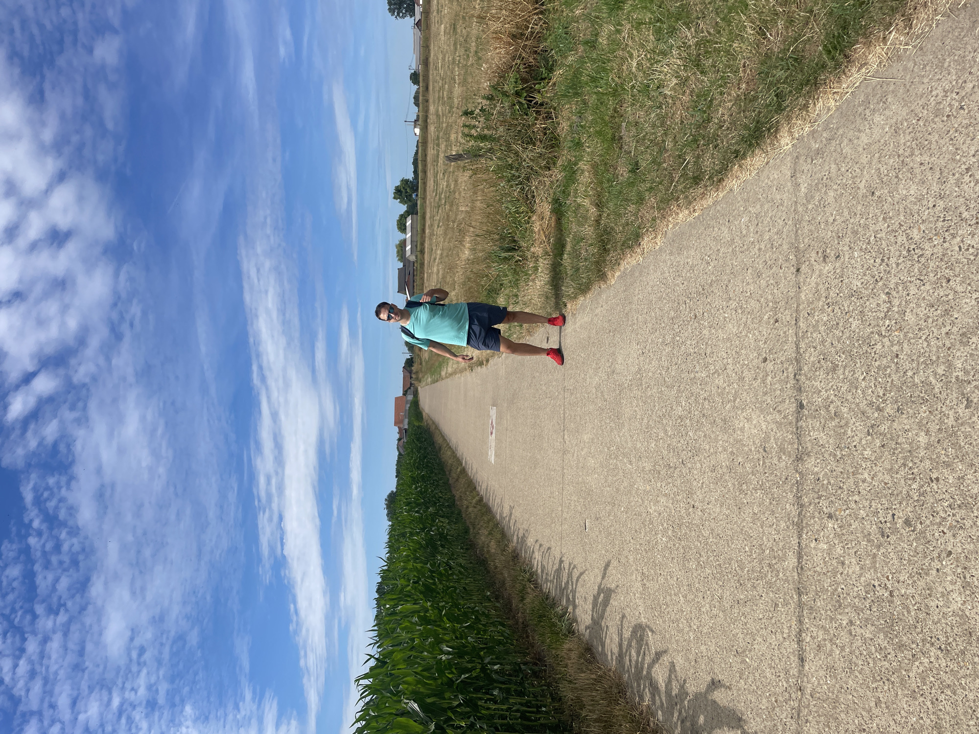 A man wearing a teal t-shirt, dark shorts, red sneakers, and a backpack stands on a concrete path in a rural area. The path is lined with a tall cornfield on the left and a dry, grassy field with scattered buildings in the distance on the right. The sky is blue with scattered white clouds. The image appears to be taken on a sunny day, likely in the summer. The location is the Kempen region of Belgium, near the village of Rooierheide in Diepenbeek.
