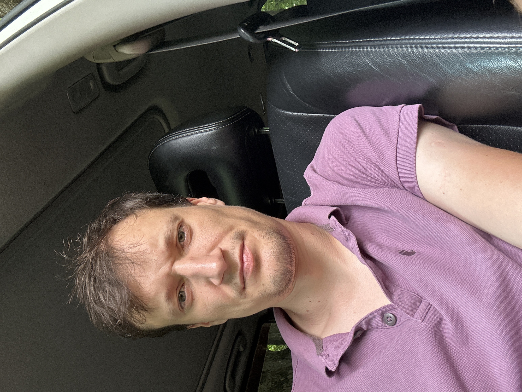 The image shows a close-up, front-facing selfie of a man in the driver's seat of a car. He has short, messy brown hair, a light beard, and is wearing a pink polo shirt. The car's interior is visible, including a black leather headrest and seat, a portion of the dashboard, and the sunroof. The background outside the car appears to be foliage, suggesting a natural or rural setting. The image quality is good, with clear focus on the man's face and upper body. There are no other people or objects of significance in the frame. The photograph was taken during the day, with natural light illuminating the scene. The location is described as Shatava, Ukraine.