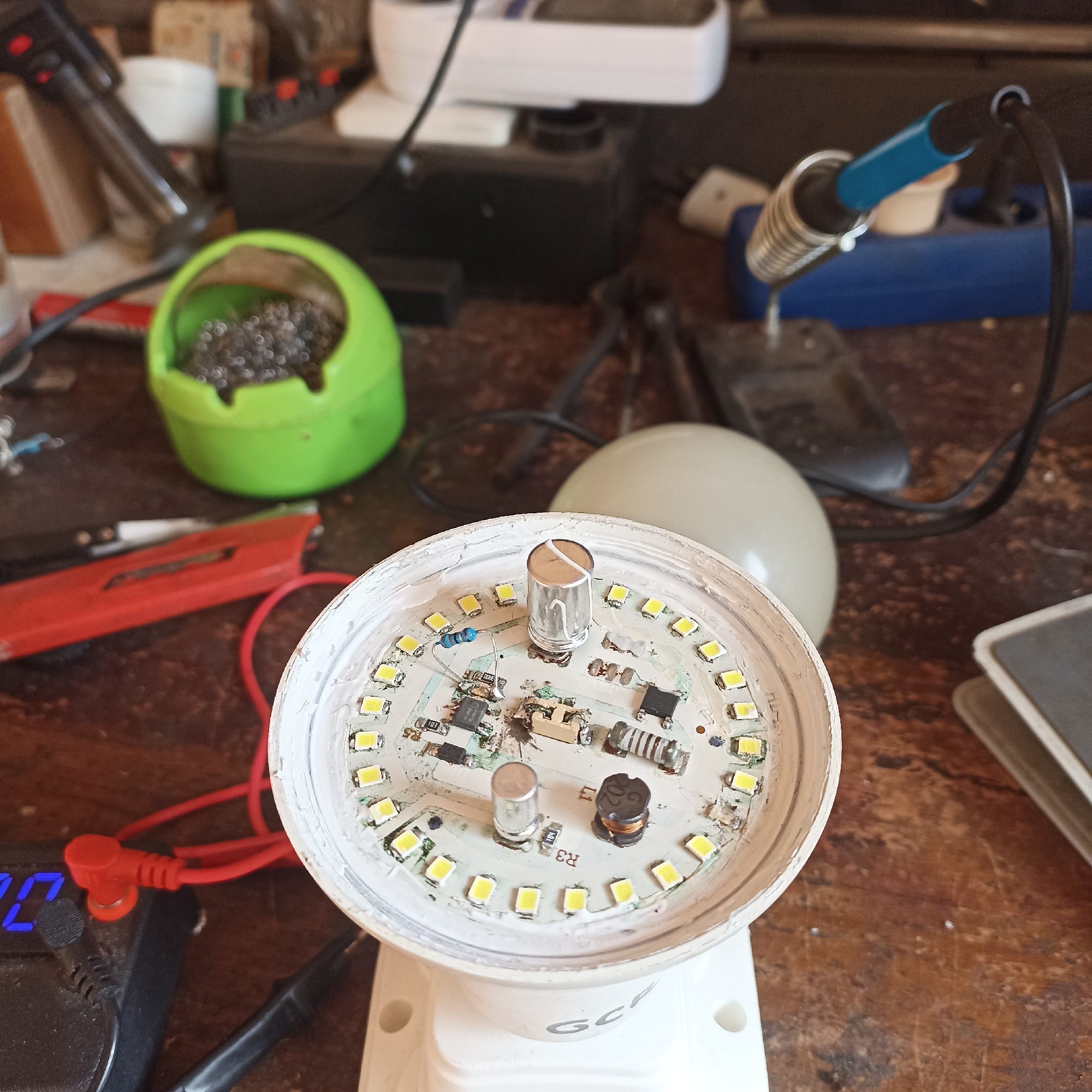 The image depicts a close-up view of a disassembled LED light bulb on a wooden workbench, likely in a repair shop or a home workshop setting. The workbench is cluttered with various electronic repair tools and components, including a soldering iron with a blue handle in a stand, a green container filled with solder, a red utility knife, a digital multimeter displaying "107" and connected with red probes, and other unidentifiable electronic parts. The LED light bulb itself has been opened, revealing its internal circuit board, which is populated with numerous small yellow LEDs arranged in a circle. The central area of the circuit board shows various electronic components such as resistors, capacitors, and what appears to be a small transformer or inductor. A shiny cylindrical component, possibly a capacitor or fuse, stands prominently in the center. White adhesive material is visible around the edges of the bulb's housing. The overall scene suggests an ongoing repair or modification of the LED light bulb. There is no indication of the specific location within the city of Magelang, Indonesia, nor any people present in the image.