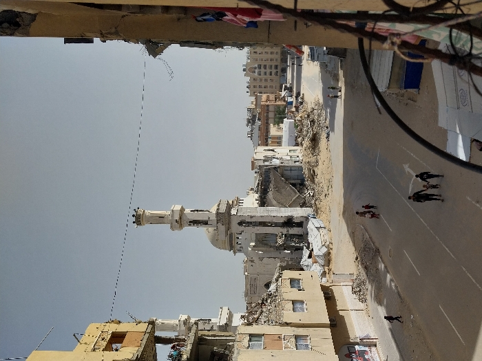 The image depicts a street scene in Khan Yunis, Palestinian Territory, observed from an elevated position. In the background, a prominent white mosque with a tall, cylindrical minaret stands significantly damaged, surrounded by large piles of rubble and debris from collapsed structures. Adjacent to the mosque, other buildings also exhibit extensive damage, including missing roofs and exposed interiors. To the left, a multi-story, light-colored building shows severe structural damage on its upper levels, with concrete partially destroyed and rebar exposed; several individuals are visible on one of these damaged floors. The foreground on the right reveals part of a damaged building facade, with an internal corner visible and what appears to be colorful laundry hanging.
The asphalt street, marked with white directional arrows and solid lines, runs through the center of the frame, bordered by sandy patches and debris on its shoulders. In the lower center, a group of four people, consisting of two adults and two children, walk hand-in-hand towards the viewer. Further up the street, to the left, another group of three children walks together. A single individual is visible walking on the left sidewalk. In the distance, several other figures are dispersed along the street. Overhead, thin, dark wires stretch horizontally across the clear, light blue sky. The overall environment suggests an urban area that has experienced significant structural destruction.
